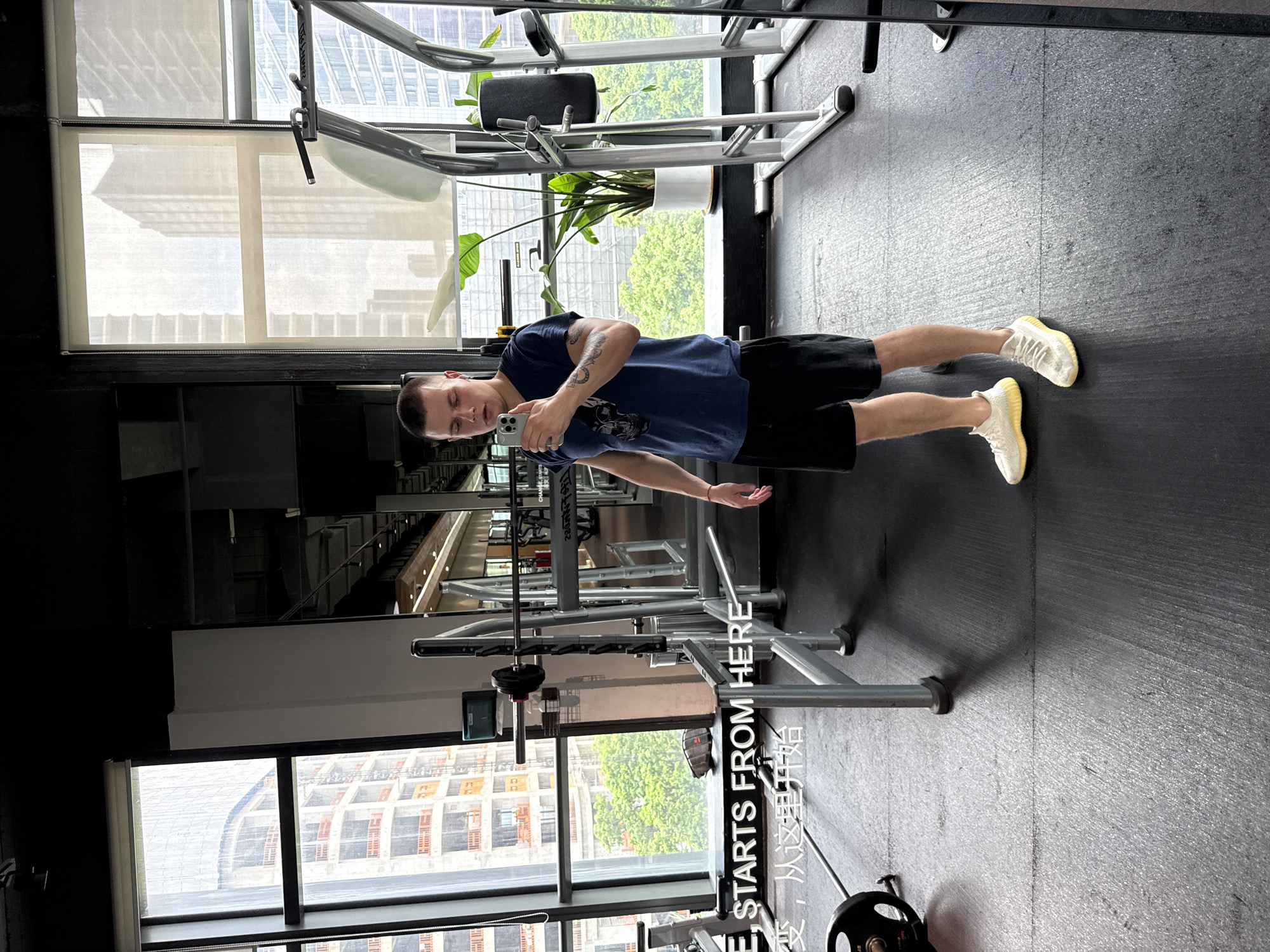 A young adult male, with short brown hair and tattoos on his left arm, stands in a modern gym. He is wearing a dark blue t-shirt with a graphic print on the front and black athletic shorts. He is holding a smartphone in his right hand, taking a mirror selfie. The gym features large windows offering a view of urban high-rise buildings, indicating its location in Haizhu District, China. In the foreground, a barbell with weights is visible on the floor, and various gym equipment, including weight racks and machines, populate the background. A large potted plant is situated near the window. The floor is a dark, textured rubber mat. Overlaid on the image is text in both English and Chinese, reading "E STARTS FROM HERE" and "变, 从这里开始".