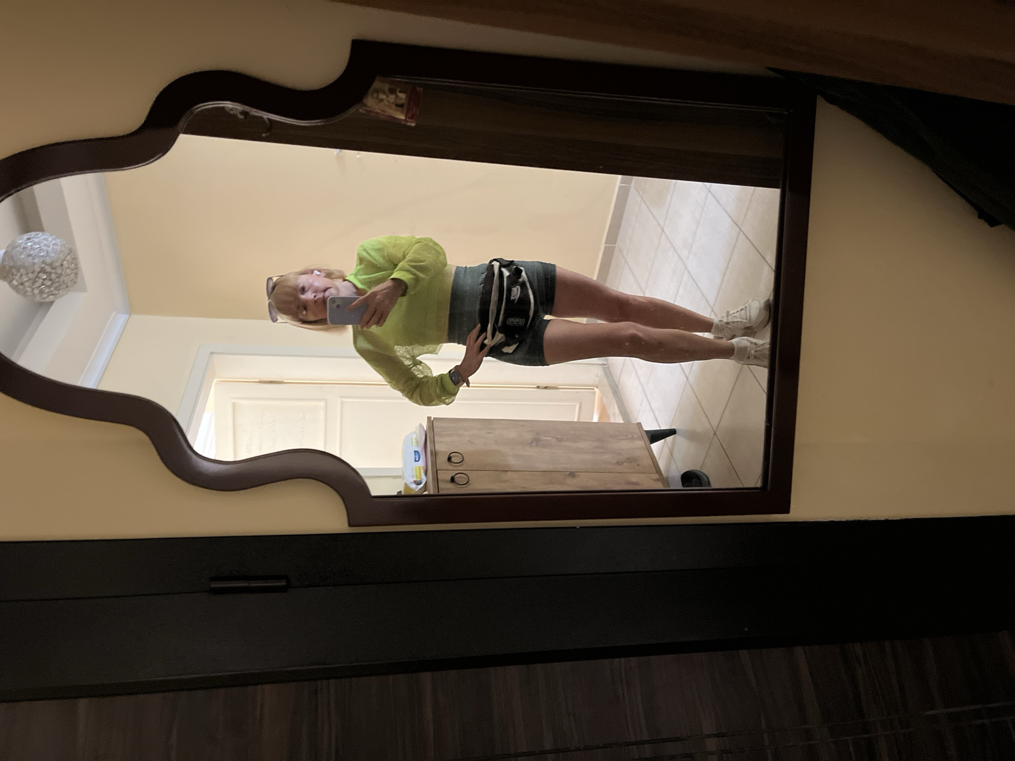 A selfie taken in a mirror in Alanya, Turkey. The mirror is ornate with a dark brown frame. A woman is reflected in the mirror, holding a cell phone and wearing a lime green mesh top, grey shorts, a black and white fanny pack, and white sneakers. Her blonde hair is shoulder length. Sunglasses are placed on her head. The mirror is mounted on a dark brown bureau. The wall is pale yellow. To the left of the mirror is a white round light fixture. The floor has beige tile. There is a partially visible wooden cabinet. A white door is in the background.