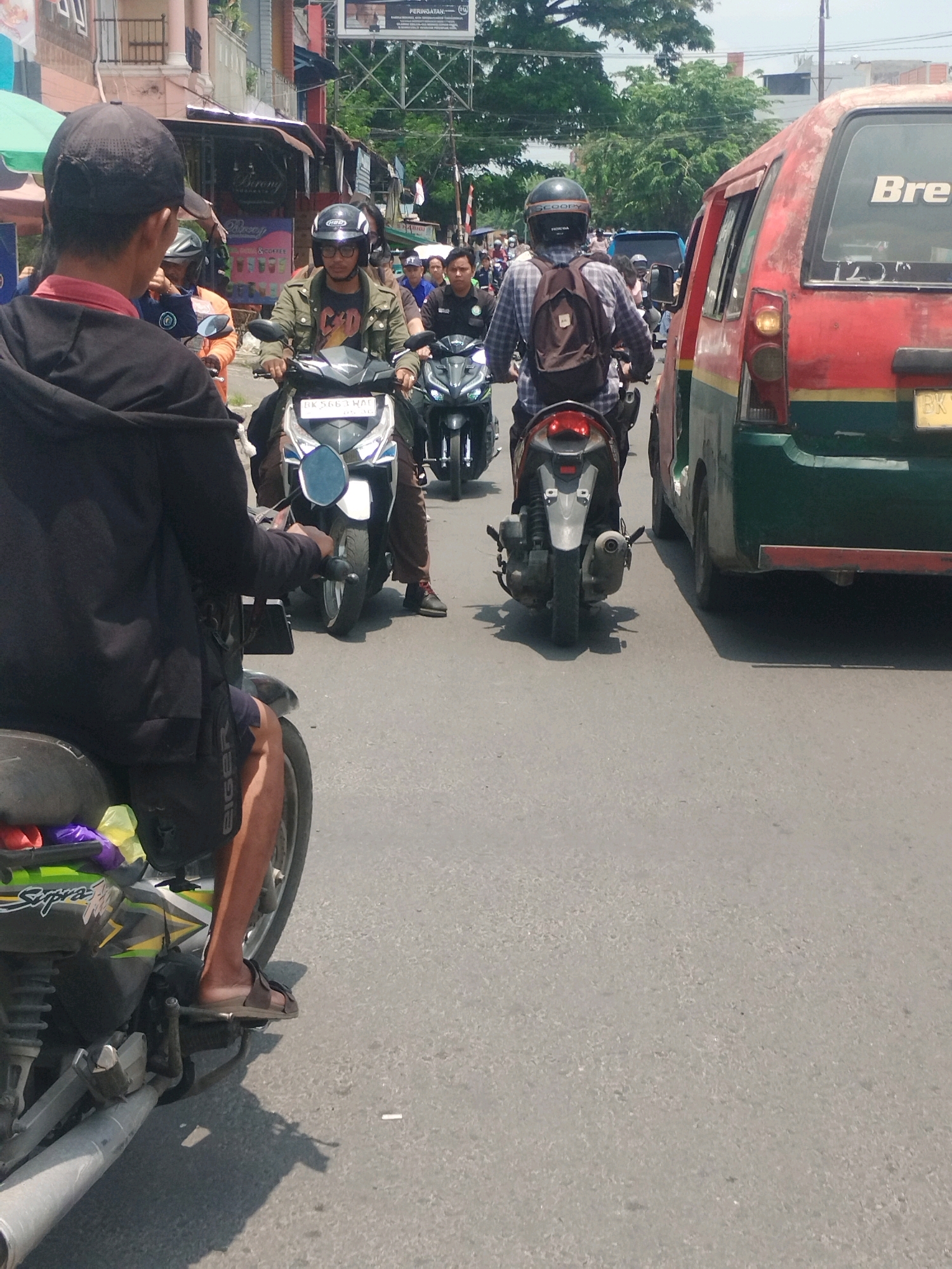 In the city of Medan, Indonesia, a bright, sunny day unfolds on a street filled with vehicular traffic. The foreground is dominated by a person on a black motorcycle, their black hoodie and black shorts visible. The motorcycle itself is a dynamic mix of black, green, and yellow, with a glimpse of a purple bag and a yellow bag secured to the rear. To the right, a faded red and green van, likely a public transport vehicle common in Medan, occupies a significant portion of the frame, with the word "Bre" partially visible on its rear.
Further down the street, a procession of motorcycles and riders creates a sense of organized chaos. One rider, wearing a green jacket and a black helmet with glasses, is on a silver and black motorcycle. Another rider, in a blue plaid shirt and a black helmet with a brown backpack, is on a white and black motorcycle. Behind them, a blue vehicle is visible, and more motorcycles and people fill the background. The scene is characteristic of bustling city streets in Indonesia, with a mix of personal vehicles and public transport navigating the roads. The asphalt is worn, and the surrounding buildings suggest a commercial or residential area.