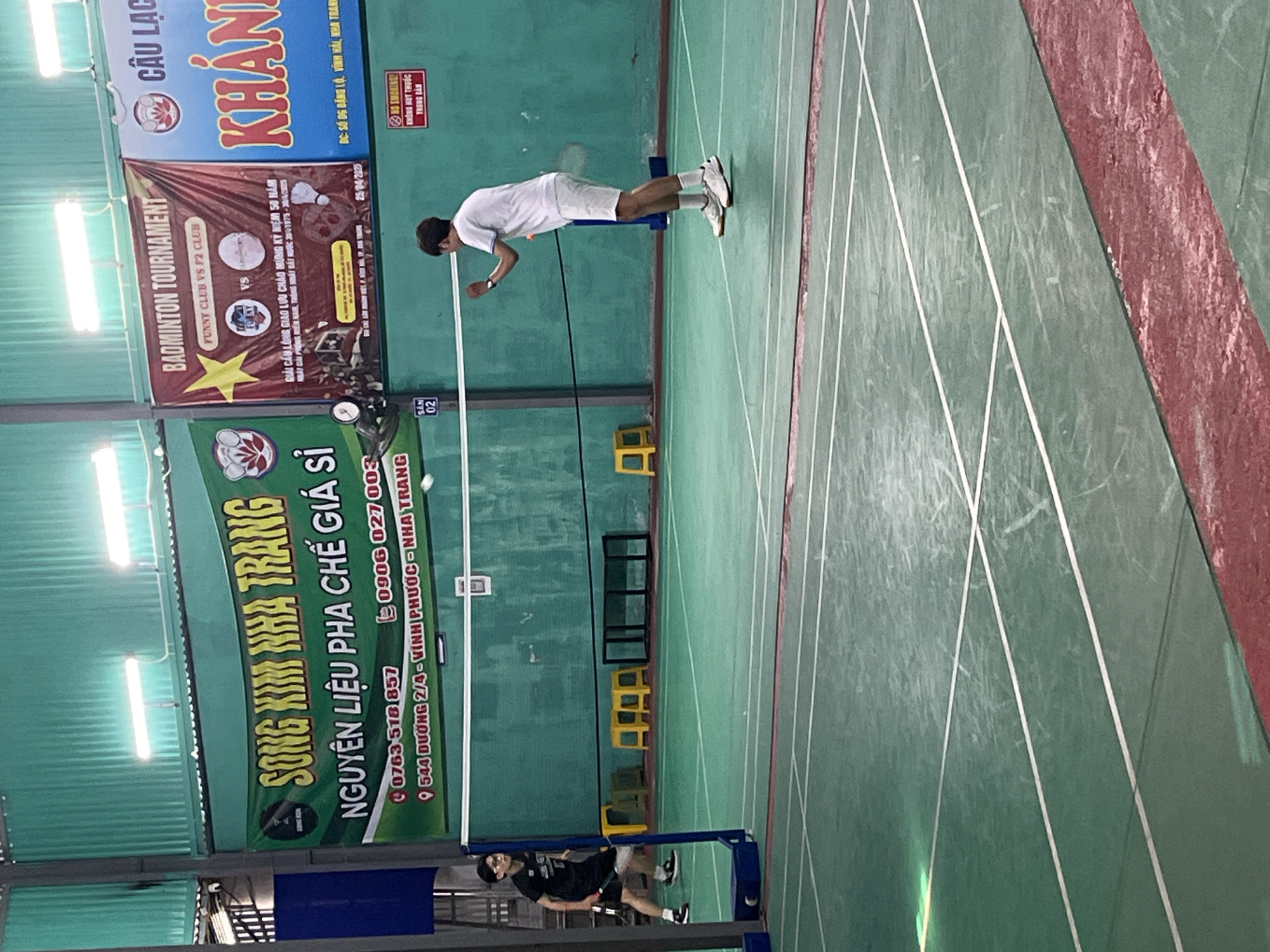 The image shows an indoor badminton court in Bac Nha Trang Ward, Vietnam. A badminton player in white clothing is positioned on the court, preparing to hit the shuttlecock. On the left side of the image is another badminton player dressed in black. Various banners and signs are hung on the walls, advertising a badminton tournament and a wholesale ingredient supplier. The badminton court floor is marked with white lines. Several yellow stools are placed on the sidelines. The image has been rotated, causing a sideways perspective.