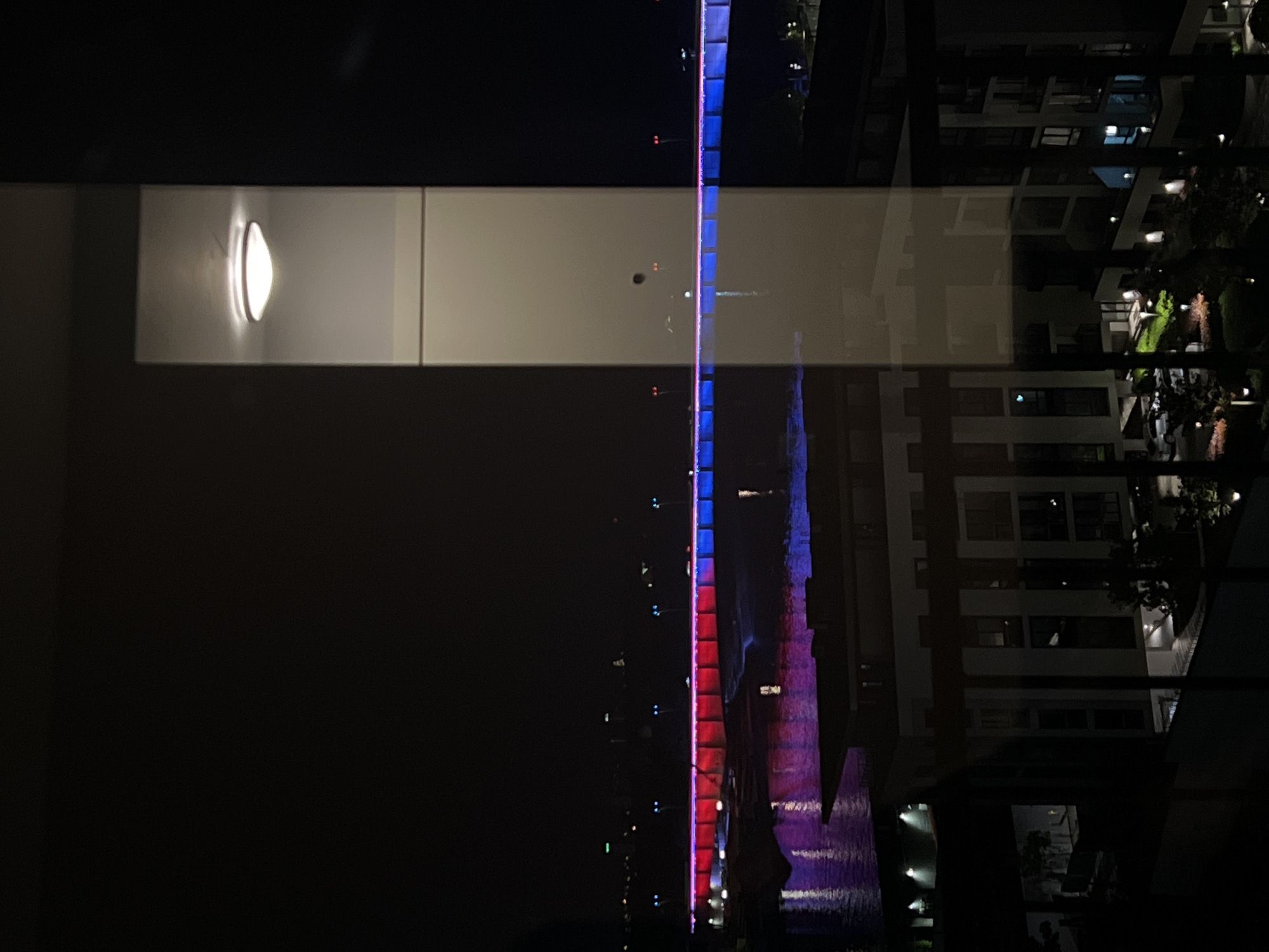 The image depicts a night scene in Belgrade, Serbia, likely taken from inside a building looking out through a window. The reflection in the window creates a layered effect, merging indoor and outdoor elements.
On the left is the interior of the room, with a softly lit wall fixture visible. The middle ground shows a bridge illuminated with red and blue lights reflecting in water below. The right side of the image reveals a partially obscured view of a building with lighted windows, some greenery, and possibly parked cars. The overall atmosphere is dark with contrasting light sources.
