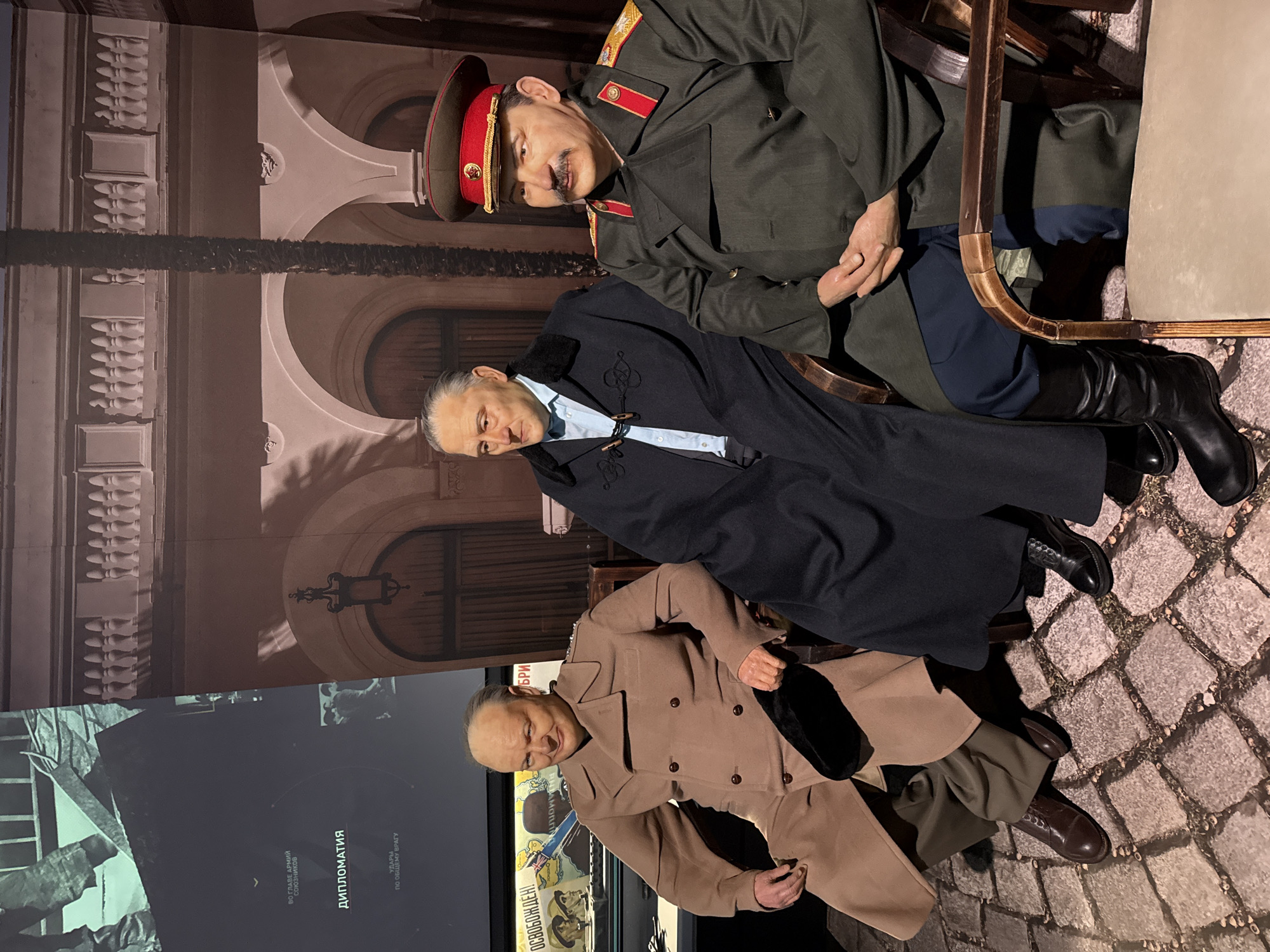 Three wax figures are seated in chairs in what appears to be a museum exhibit. The figure on the left is Winston Churchill, recognizable by his bald head and jowly face, wearing a tan trench coat and holding a black hat in his lap. The figure in the center is a man with graying hair, wearing a dark blue overcoat with fur trim over a light blue shirt. The figure on the right is Joseph Stalin, wearing a military uniform with a red cap and high black boots. He has a mustache and is depicted with his hands clasped in his lap. The figures are seated on a cobblestone-patterned floor. Behind them is a mural depicting an arched building with palm trees, and to the left, a large screen displays images and text, including "ДИПЛОМАТИЯ" (Diplomacy). The exhibit is located in the city of Zelenaya Roshcha, Russia.