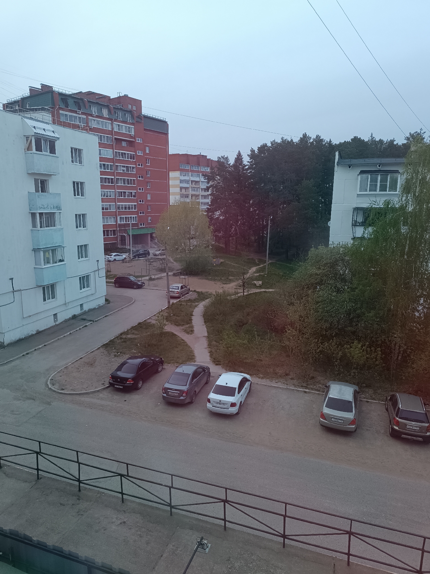 The image depicts a view from an elevated position, likely a balcony, overlooking a residential courtyard in what appears to be Chaikovsky, Russia. Several multi-story apartment buildings are visible in the background. The buildings are constructed of concrete and brick, painted in white, red, and beige. A pathway meanders through a green space with trees and shrubs. In the foreground, several cars are parked along a paved area adjacent to the path. The cars are various colors, including black, gray, and white. A black metal railing runs along the bottom of the frame, suggesting the viewpoint is from a balcony or window. The sky is overcast, suggesting an overcast day. No people are visible in the image.
