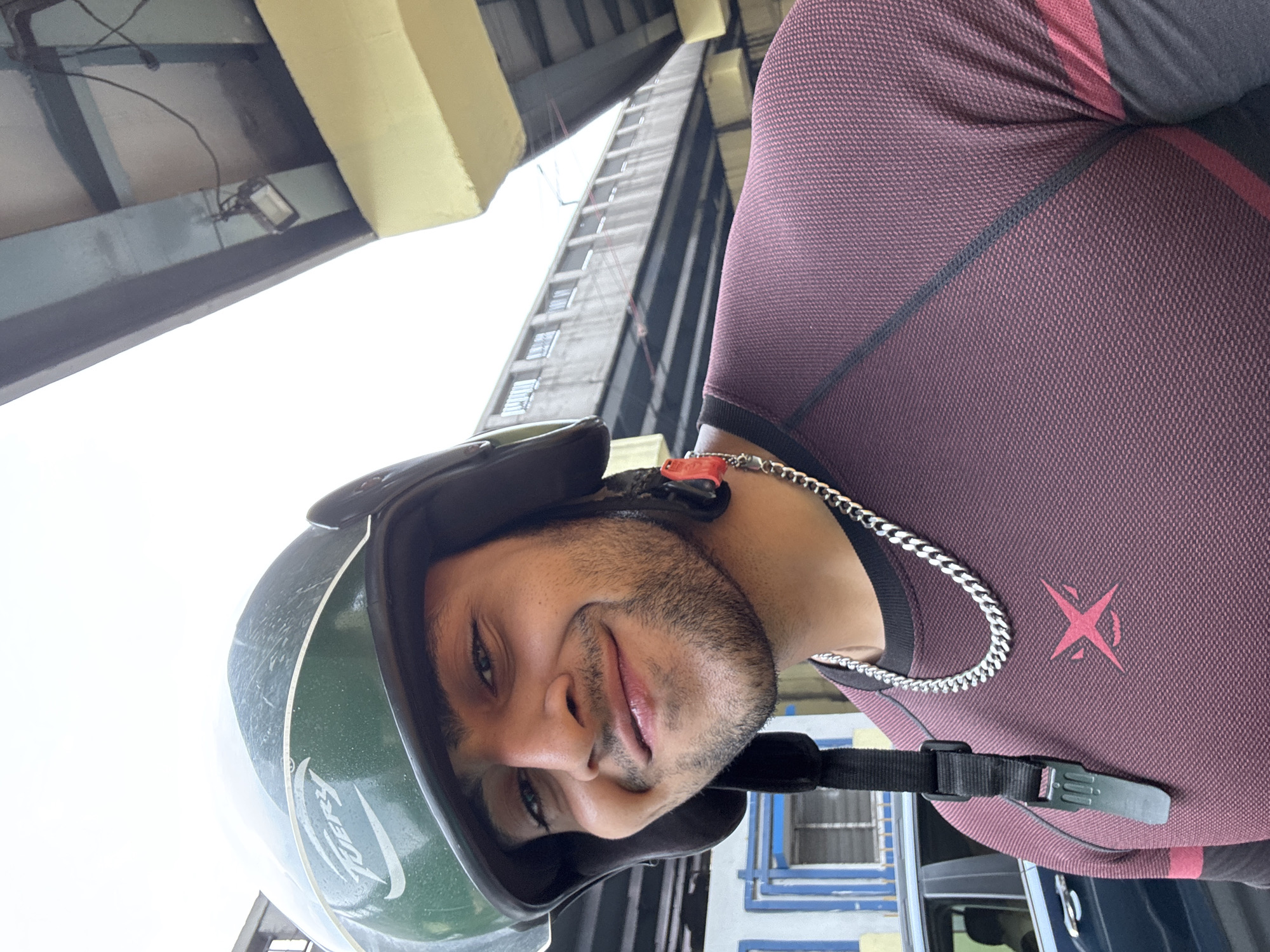 A young man wearing a dark green helmet and a maroon long-sleeved shirt with a silver chain is smiling at the camera. He has a short beard and appears to be outdoors in Kolkata, India. The background shows a yellow overpass and a blue building.