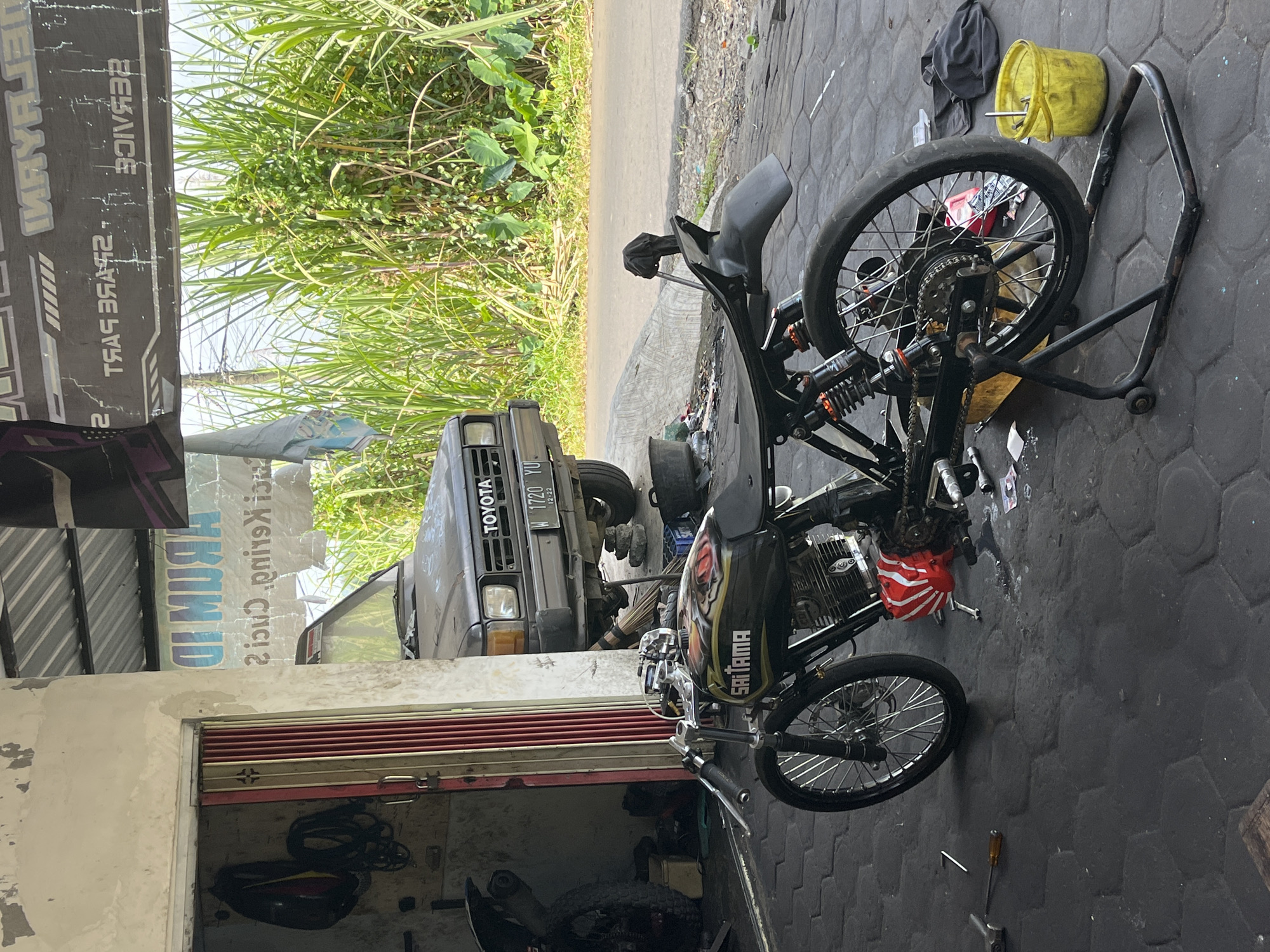 The image depicts an outdoor motorcycle repair shop in Pasrukkrajan Satu, Indonesia. A custom motorcycle, painted black with red and gold accents and the word "SAITAMA" on the fuel tank, is parked on a stand in the foreground. Behind it, a dark-colored Toyota SUV is partially visible, along with a rolling metal door of the shop. The ground is paved with dark hexagonal tiles. A tall, dense growth of green vegetation, possibly sugar cane, borders the scene. The overall environment suggests a functional, working space with tools and equipment scattered around.