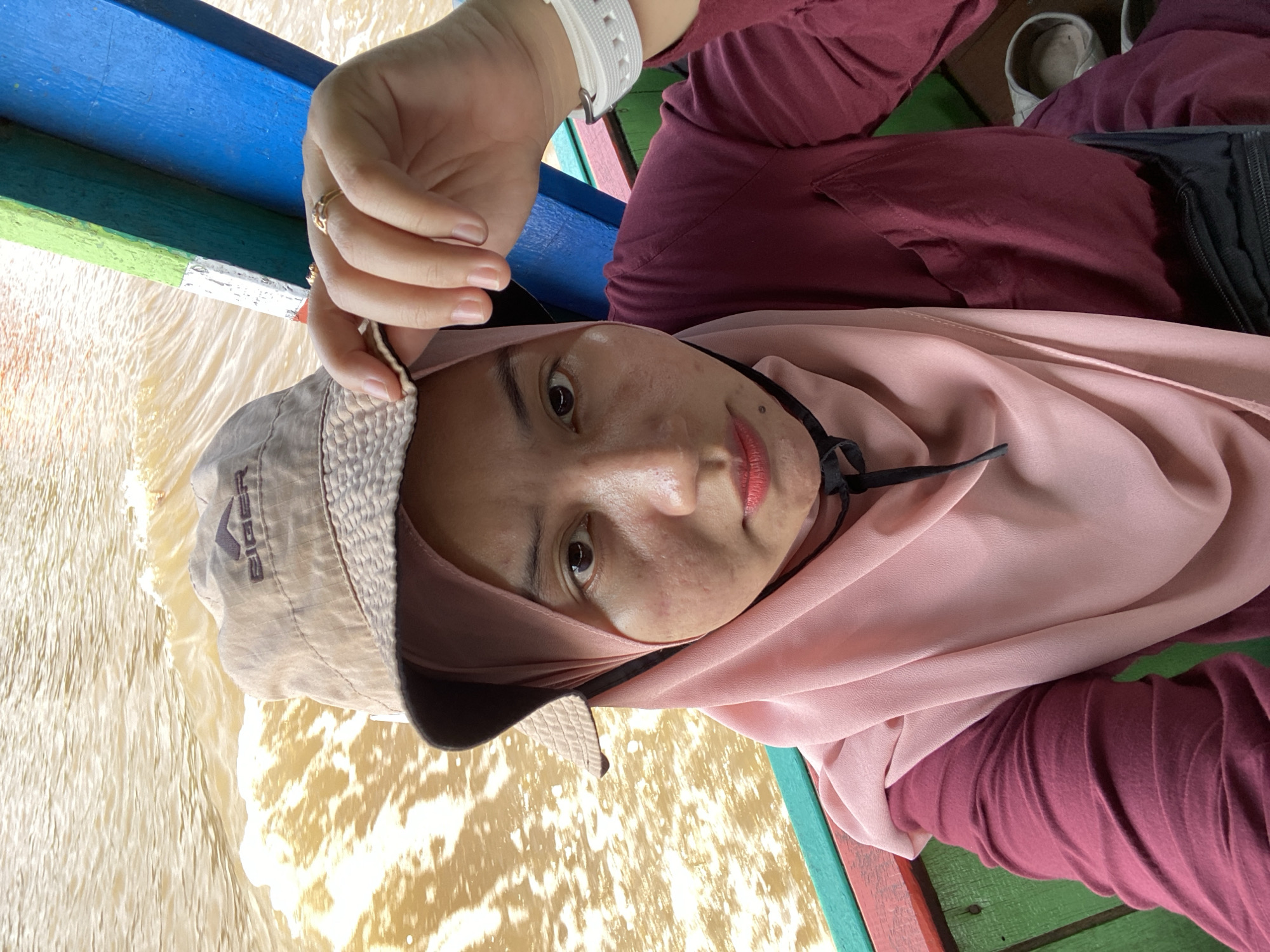 A selfie shows a woman wearing a light brown bucket hat with the "Eiger" logo, a pink hijab, and a maroon long-sleeved shirt. She is seated on a wooden boat with green, red, and blue painted railings, on murky brown water. The woman is touching the brim of her hat with her right hand, on which she wears two gold rings. Her left wrist has a white watch. The background is water with visible ripples and foam. The photo was taken outdoors, presumably in Berangas Timur, Indonesia, given the context.