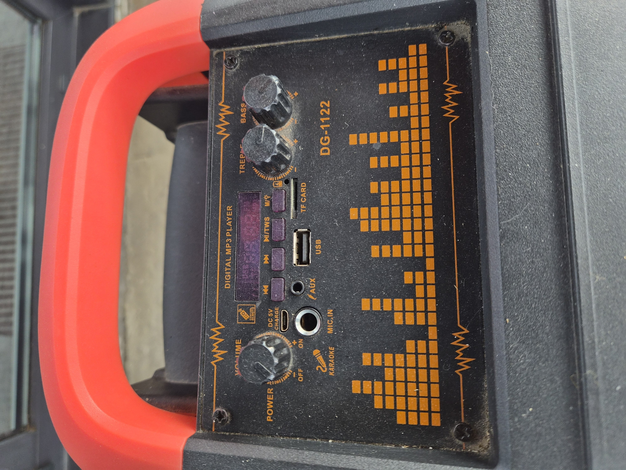 This is a close-up, eye-level, outdoor shot of a portable speaker with a bright orange handle. The speaker is black and rectangular, with a prominent equalizer display made of orange squares on the right side of its control panel. The control panel also features several knobs, buttons, and input ports.
Two knobs are labeled "TREBLE" and "BASS." Below these, a small digital screen is visible, along with buttons labeled "MODE," "M/TWS," and playback control symbols. Further down, there are ports for "TF CARD," "USB," and "AUX." Below the "USB" port, there is a "DC 5V CHARGE" input, a microphone input labeled "MIC.IN," and a switch labeled "POWER" with "OFF," "ON," and a "+" and "-" indicator. A button labeled "KARAOKE" is also present.
The speaker appears to be dusty, suggesting it might be used outdoors or has been stored for some time. The bright orange handle curves around the left side of the speaker. The background is blurred, but appears to be a gray, textured surface, possibly a wall or pavement. There are no people or animals visible in the image. The overall impression is that of a functional, though somewhat weathered, piece of audio equipment.