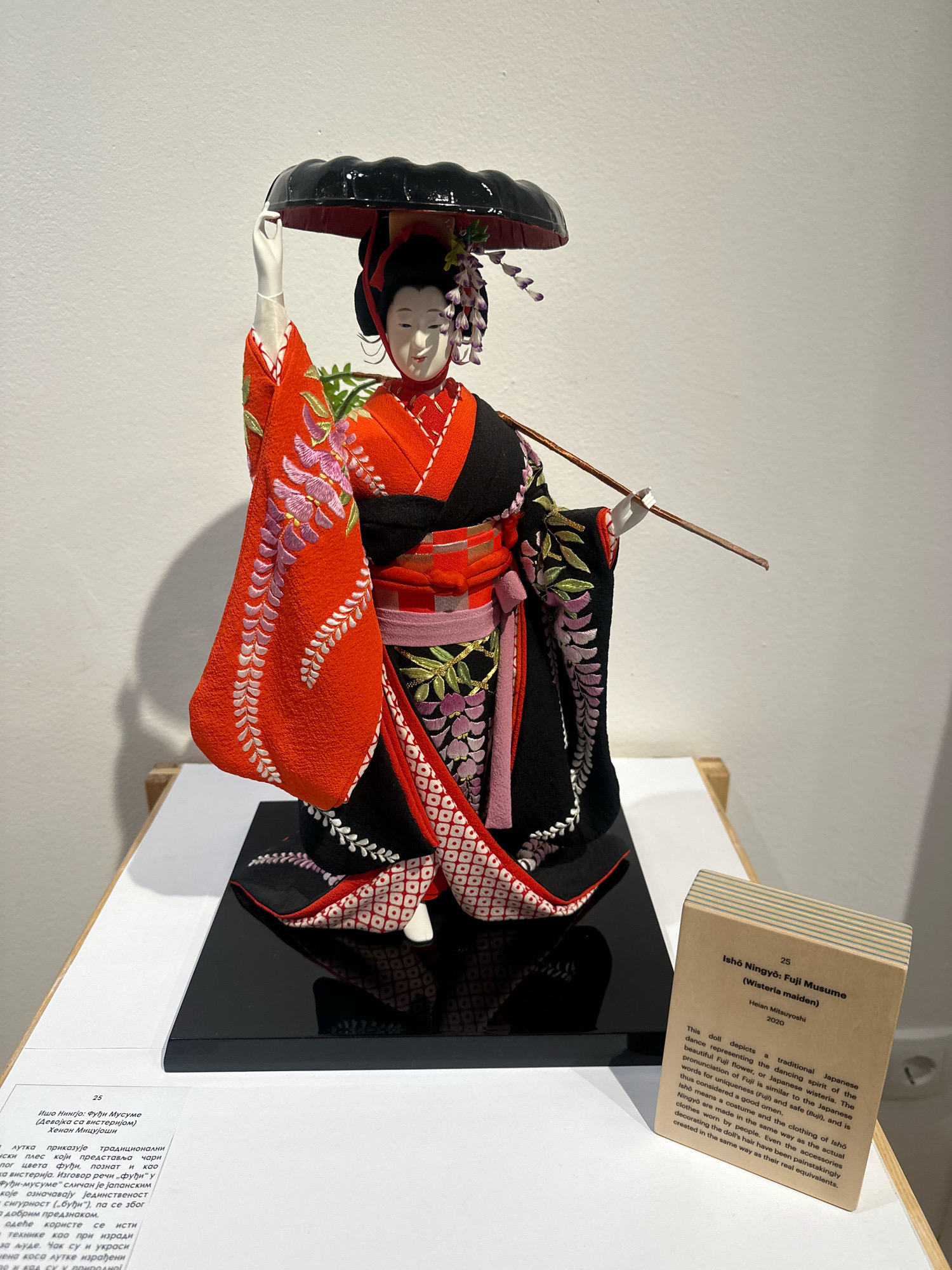 This image depicts a traditional Japanese doll, identified by a plaque as "Ishō Ningyō: Fuji Musume (Wisteria maiden)" by Heian Mitsuyoshi, dated 2020. The doll is dressed in a vibrant orange and black kimono adorned with floral embroidery, holding a black umbrella above its head and a stick-like object. It is displayed on a black base against a plain white wall. To the right of the doll, a beige information card provides details about the artwork in English. Below the doll and to the left, another information card, partially visible and with text in a Cyrillic alphabet, is also present. The overall presentation suggests an exhibition or museum setting. The presence of Cyrillic text strongly indicates the location is in a city where this script is used, such as Belgrade, Serbia. The lighting suggests an indoor environment with overhead illumination casting shadows on the white wall.