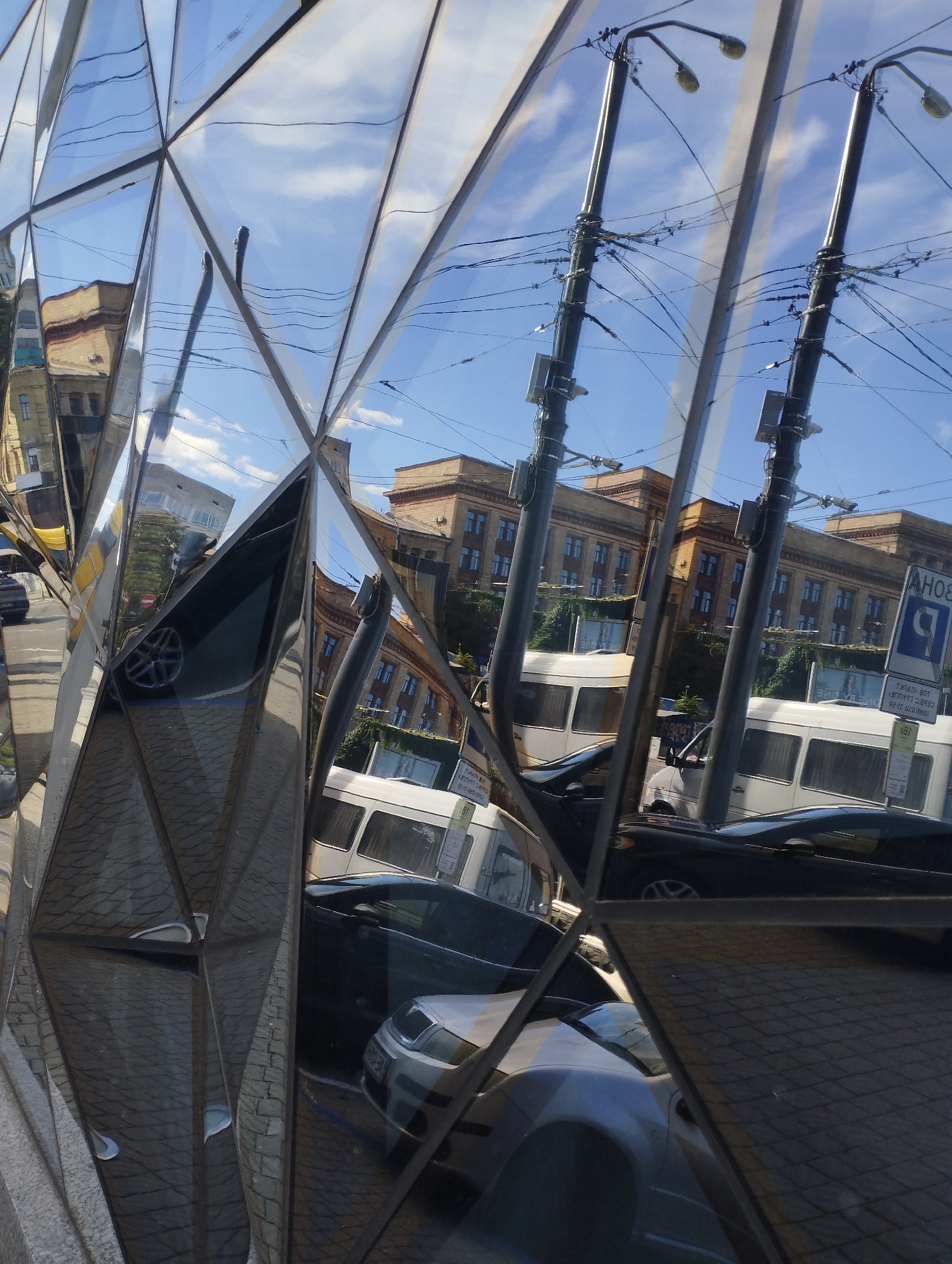 The image is a reflection on a faceted, mirrored surface, showing a street scene. The reflections include buildings, utility poles with wires, cars, and a blue sky with clouds. One building appears to be light brown with multiple windows. There are at least two white vans and several cars visible, including a dark sedan and a lighter colored car. A blue and white parking sign is also reflected. The mirrored surface distorts the scene, creating geometric patterns. The image is taken from a low angle, looking up at the reflected scene. The location is described as Unknown, Unknown.
