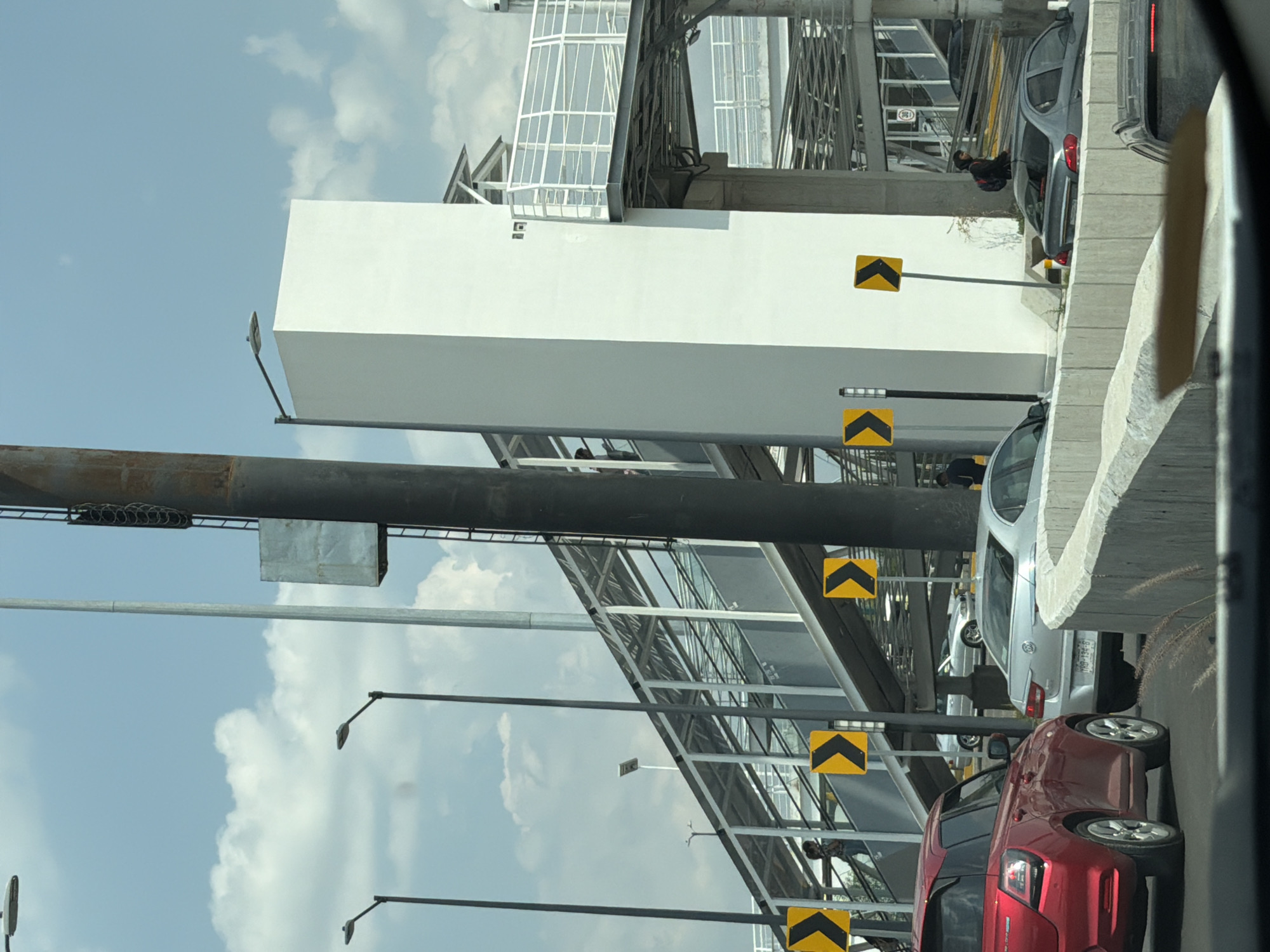 The image shows a traffic scene in San Bernardino Tlaxcalancingo, Mexico, taken from inside a vehicle. Several cars are visible on the right side of the image, including a red car, a silver car, and a grey car. The cars are separated by a concrete barrier. Overhead, a large metal structure spans the road with white panels and metal framework. Below the structure, multiple yellow chevron signs are attached to poles. The sky is blue with scattered clouds. Streetlights are also visible. People appear to be standing underneath the metal structure, near where the cars are stopped.