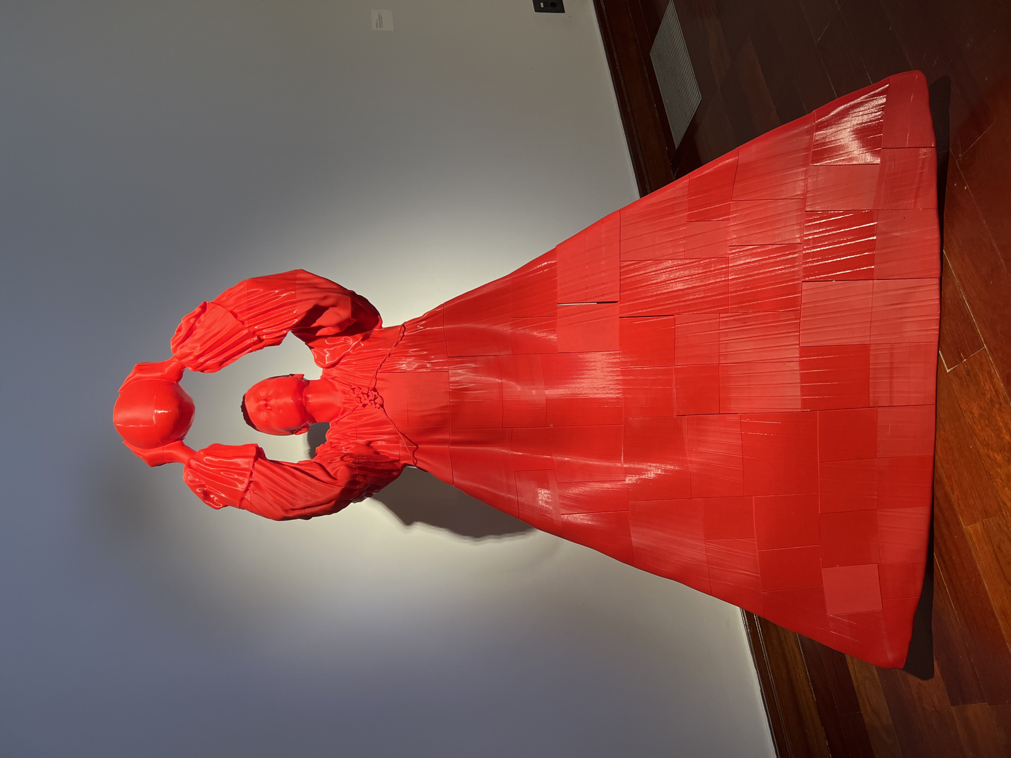 A red, modern art sculpture of a person in a long gown, holding two spherical objects above their head, stands on a hardwood floor against a gray wall. The sculpture appears to be constructed from numerous rectangular components. The location is Las Palmas, Spain, based on available metadata.