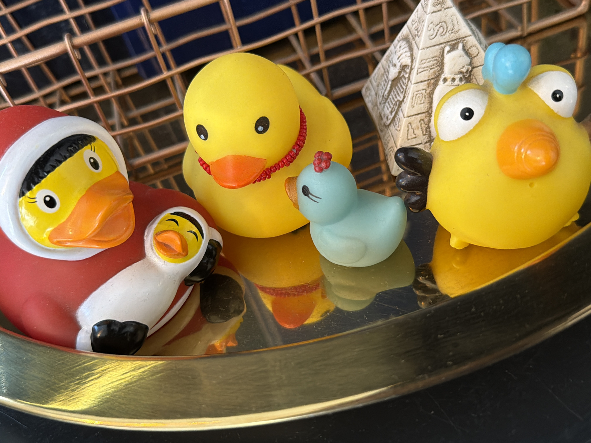The image features a collection of rubber ducks and a small pyramid-shaped object arranged on a reflective gold-colored tray. The rubber ducks include a standard yellow duck, a blue duck with a red flower on its head, a yellow chick with a blue tuft, and a duck dressed in a red and white Santa Claus outfit with a smaller duck in a similar outfit nestled against it. The pyramid is light-colored with Egyptian-style figures on its sides. The tray is placed on a dark surface, likely a table, and the background includes a wire basket with a dark blue interior.