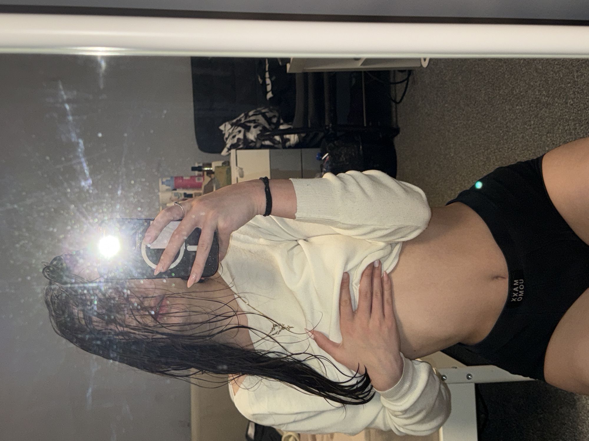 A young woman is taking a selfie in front of a mirror. She is wearing a white sweater and black shorts. Her hair is wet. The location is not identifiable as Sofia, Bulgaria, based on the image provided. The background includes a desk, bag and other objects.