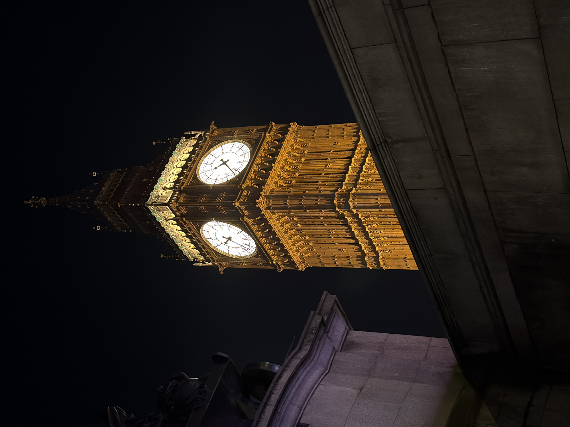 The image depicts the Elizabeth Tower, part of the Palace of Westminster (Houses of Parliament) in London, United Kingdom, at night. The tower is brightly illuminated, and two clock faces are visible, displaying different times. The lower portion of the frame shows other architectural elements of the building, constructed from stone. The sky is dark, suggesting nighttime.