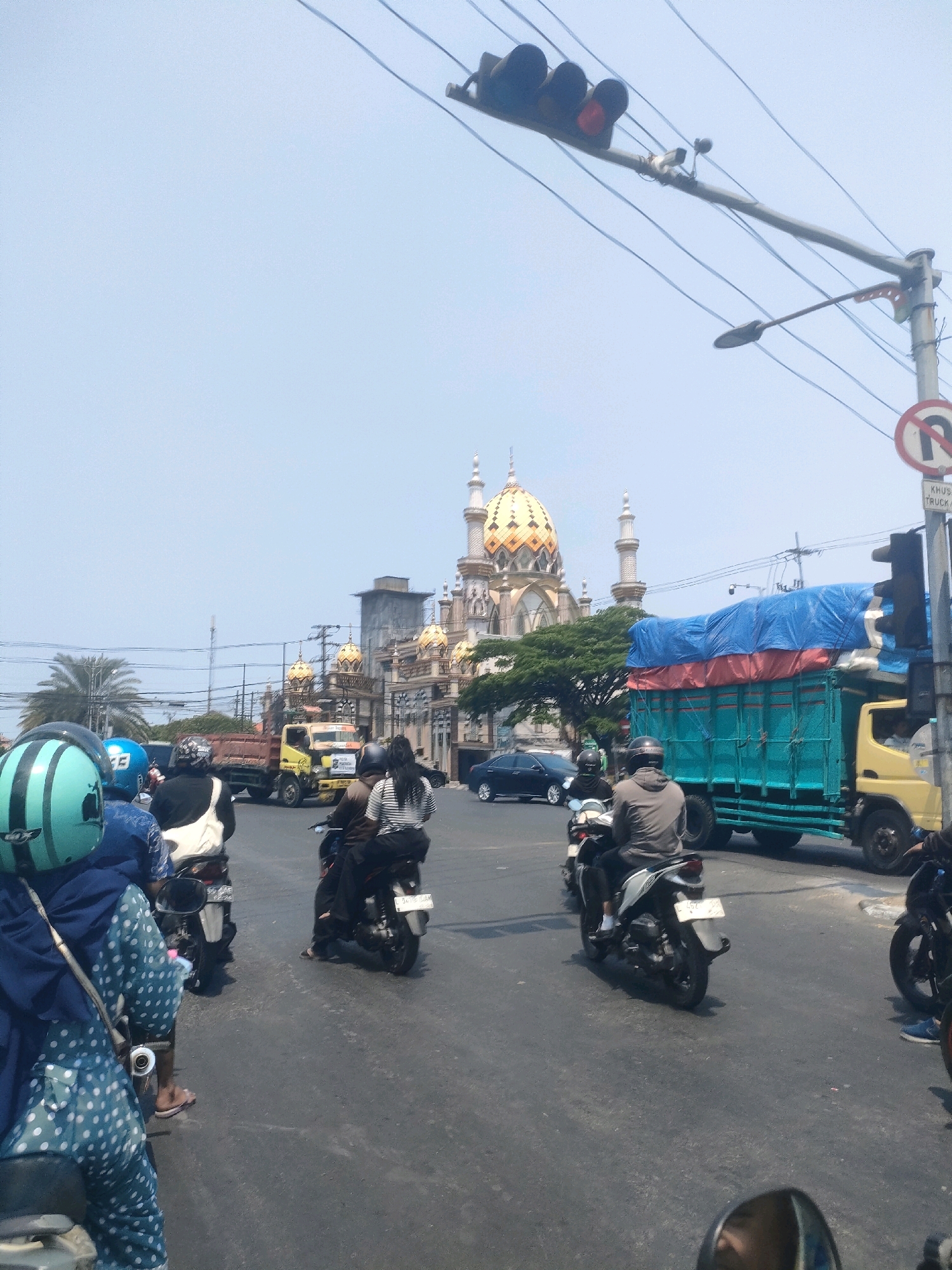 This is a street-level, eye-level, medium shot photo taken in Surabaya, Indonesia. The scene captures traffic on a sunny day, with a prominent mosque in the background. In the foreground, several people are riding motorcycles. One person in the left foreground is wearing a light blue helmet with green stripes and a dark blue patterned outfit. Next to them is another person wearing a white jacket. In the middle ground, a yellow truck with a blue tarp-covered load is visible, along with a turquoise truck. Behind the trucks, a mosque with a golden dome and minarets stands out against the clear blue sky. Traffic lights and power lines are also present, indicating an urban environment. The road surface is asphalt and appears to be somewhat worn.