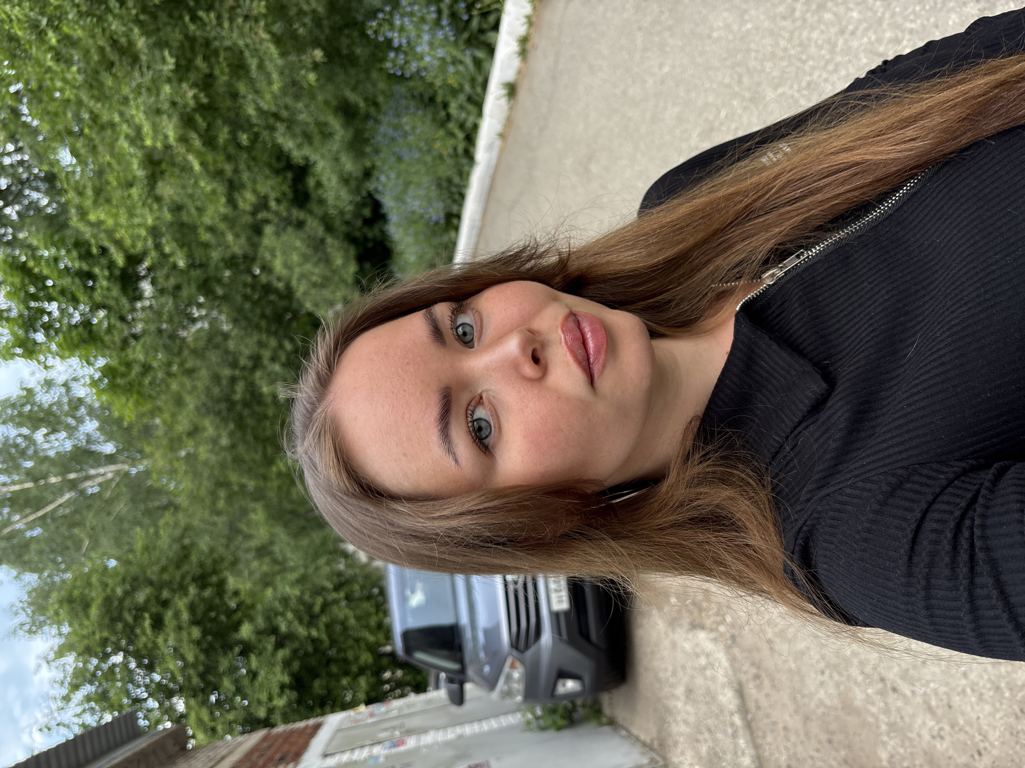 A young woman with light brown hair and blue eyes is posing for a selfie outdoors. She is wearing a black ribbed jacket with a silver zipper. Behind her, a gray van is parked. The background includes greenery and a building. Based on the plate on the car, this image was taken in Izhevsk, Russia.