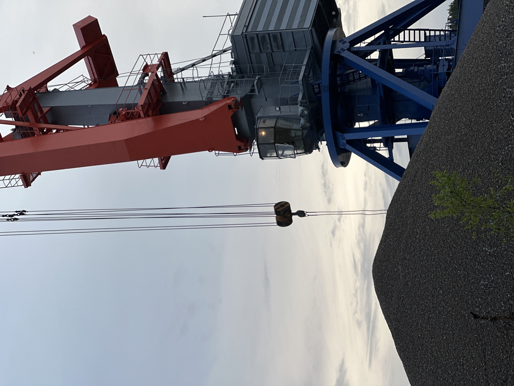 The image shows a large crane at what appears to be an industrial site in Chaikovsky, Russia. The crane is red, gray and blue. A large pile of what looks like gravel or asphalt aggregate is visible to the lower right of the crane's base. The sky is overcast. The crane's hoist cable is extended, and the hook is suspended. No people are visible.