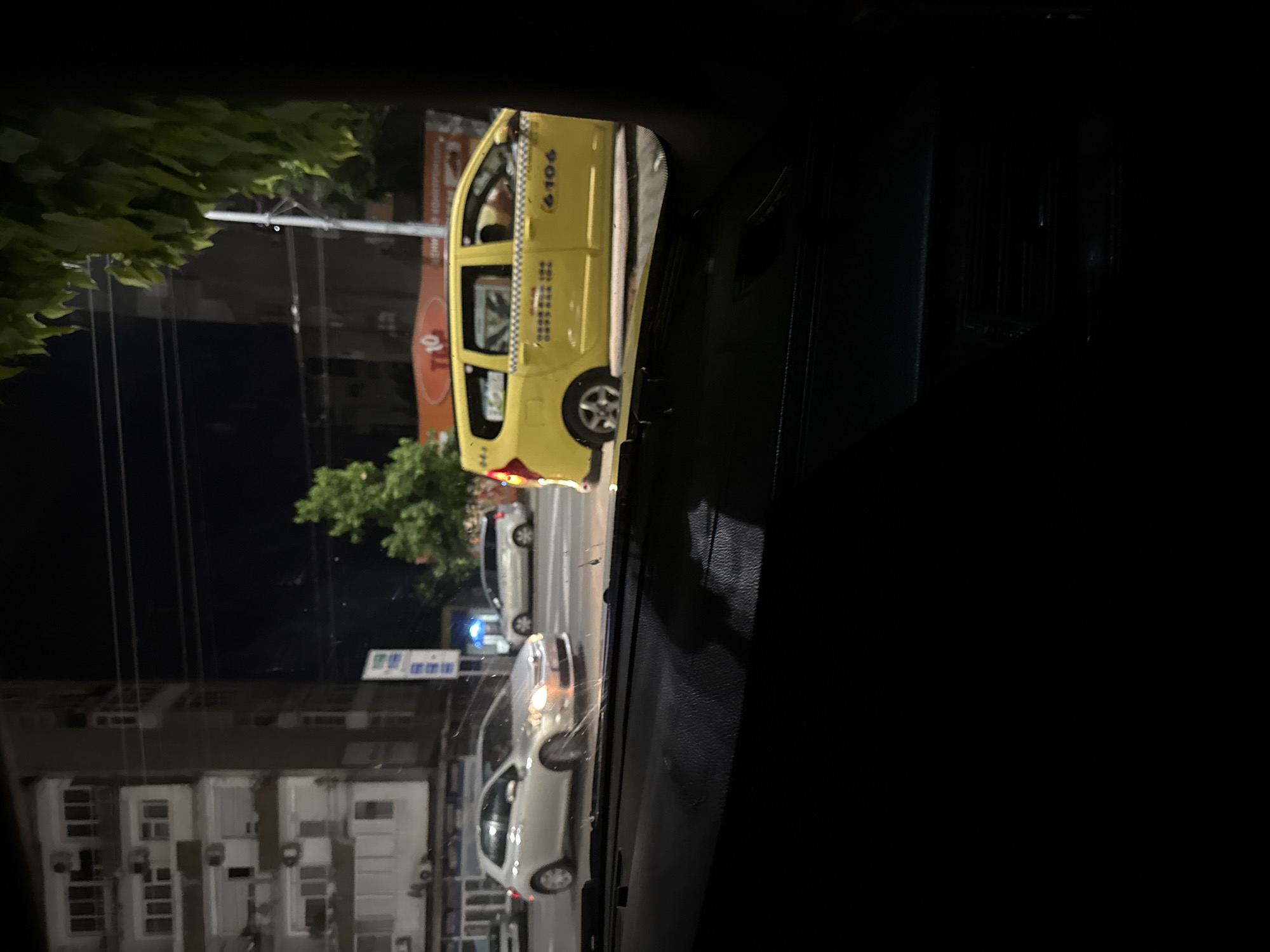 Viewed from inside a vehicle at night, a street scene in Stara Zagora, Bulgaria, is depicted. The immediate foreground shows the dark dashboard and lower windshield. Beyond, the urban environment is illuminated by artificial lights against a dark sky.
On the left side of the street, a multi-story residential building stands, featuring numerous balconies, external air conditioning units, and roller shutters on its windows. Several parked vehicles are visible along the curb in front of this building.
In the midground, a silver five-door hatchback, resembling a Toyota Auris, moves from left to right with its headlights illuminated. To its right, a yellow Dacia Logan MCV taxi is partially visible, facing right. The taxi displays the number "6106" and a Bulgarian mobile phone number starting with "089" on its side, along with a black-and-white checkered strip. Further down the street in the background, a white minivan is also visible.
Overhead utility lines crisscross the dark sky on the right side of the image, partially obscured by green tree foliage in the upper right. A tall, vertical blue sign, possibly indicating a gas station or commercial complex, stands roadside. Below this, an orange horizontal sign displaying "T 10" is clearly visible, identifying it as a "TI 10" convenience store. The scene portrays a bustling commercial and residential thoroughfare at night.