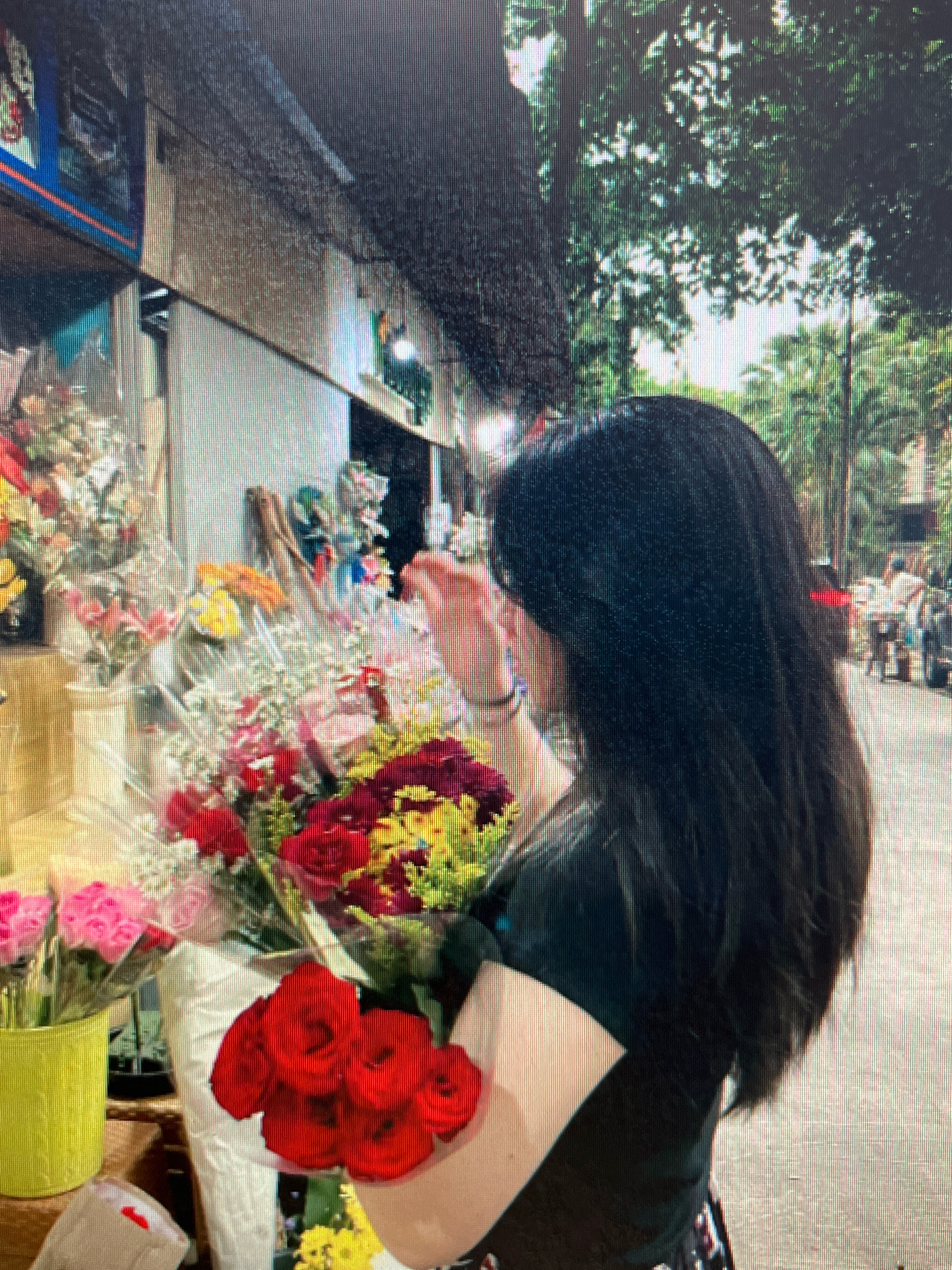 A woman with long, dark hair is seen from the side, standing in front of a flower stall. She is wearing a dark top. She is holding a bouquet of red roses and appears to be looking at or arranging the flowers. The flower stall is filled with various colorful bouquets, including pink roses in a yellow pot, and other red and yellow flowers. The background shows a street scene with trees and other people, suggesting an outdoor market or sidewalk setting. The image has a slightly blurry and pixelated quality. The location is identified as the city of Sejahtera, Indonesia.
