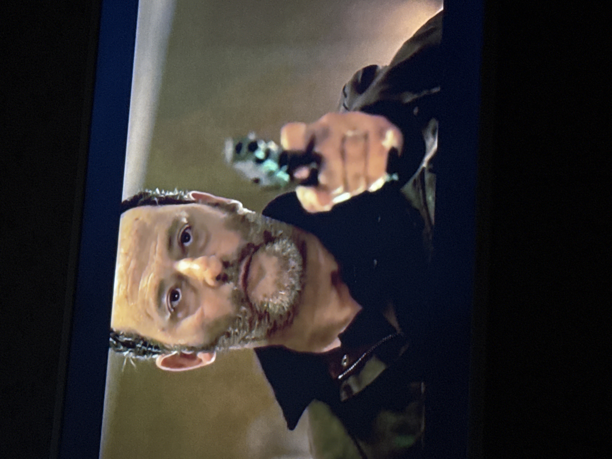 The image shows a man pointing a gun. The man is white with short, dark hair and a salt-and-pepper beard. He is wearing a dark jacket. The background is blurry and indistinct. There is no visible indication of the city of Mytishchi, Russia.