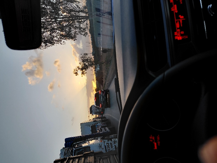 Inside a car, the view is towards a sunset sky with scattered clouds. A line of vehicles, including trucks, waits on the road. The car's dashboard and steering wheel are visible, with illuminated red displays on both. A rearview mirror occupies the top left corner. The scene suggests the tail end of a traffic jam at dusk.