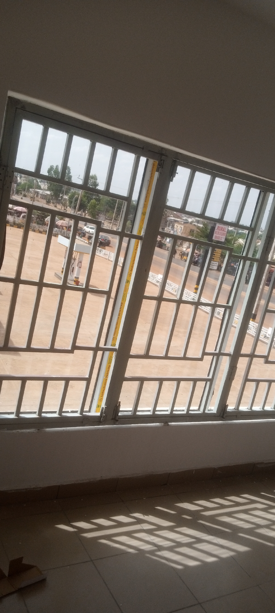 A window with metal bars looks out onto a scene in Sabon Tasha, Nigeria. A gas station is visible outside, with cars and motorcycles driving on the road. A yellow measuring stick is attached to the window frame. Shadows from the window bars are visible on the floor inside. A sign that says "RURAL" is displayed on a store near the gas station.