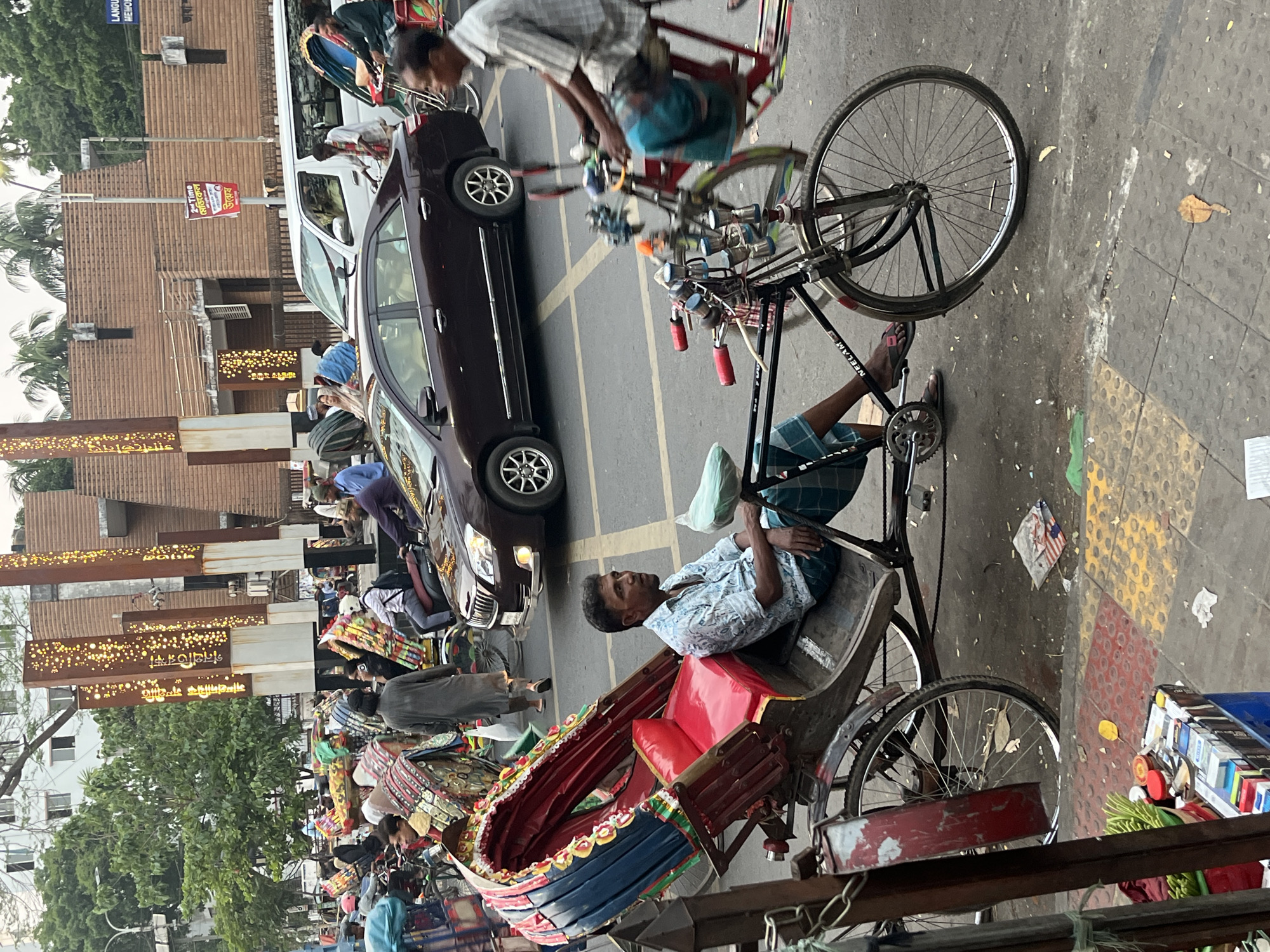 The image depicts a street scene in Dhaka, Bangladesh, featuring a parked dark brown car alongside a row of rickshaws and their drivers. One rickshaw driver is sitting in his vehicle. Another is operating his rickshaw in the background. There are many pedestrians, some with bags, and signs are visible in the background.