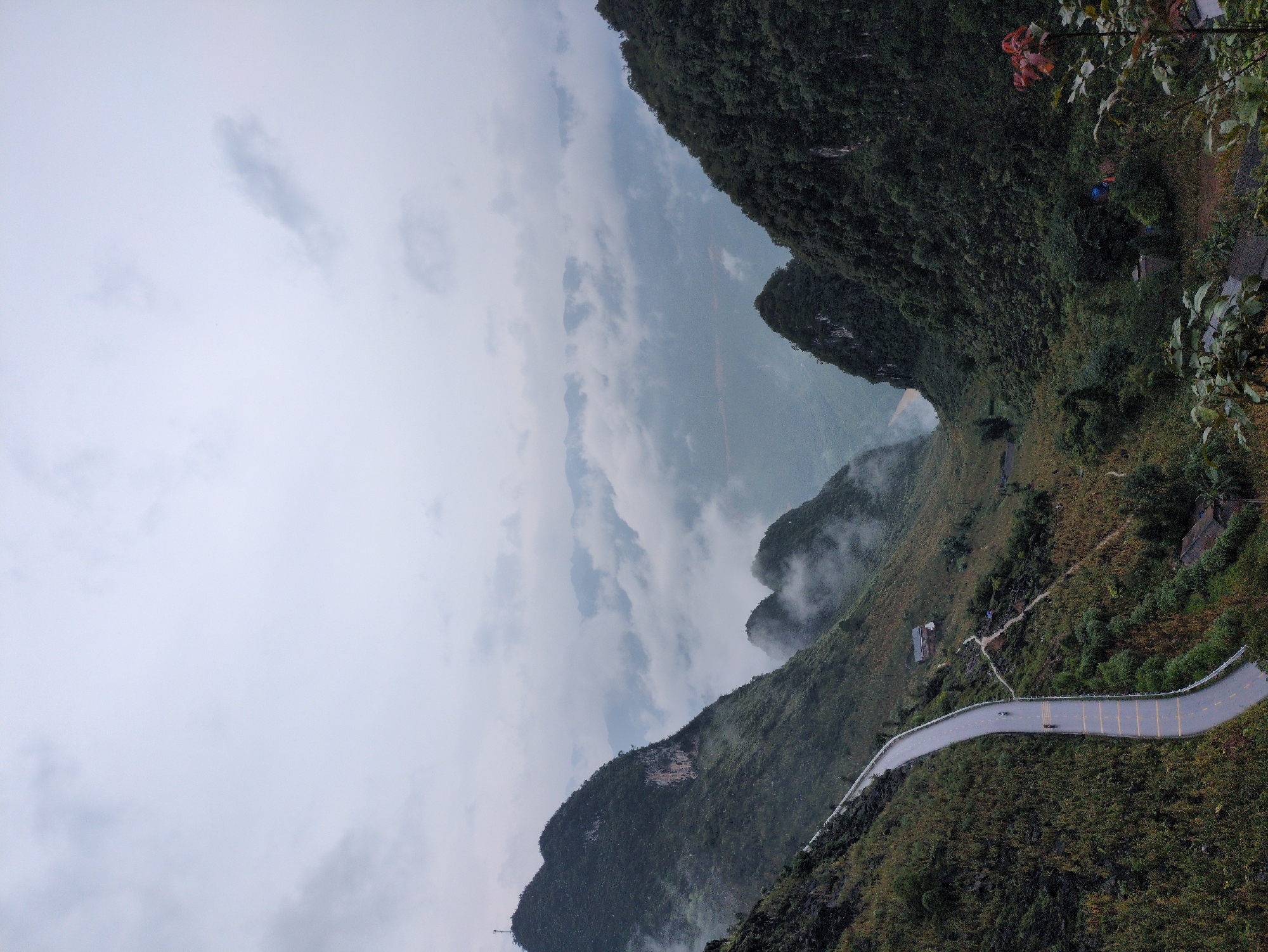 The image depicts a scenic mountain landscape under an overcast sky, likely taken on a cloudy or foggy day. In the foreground, a winding asphalt road descends from the top left of the frame, curving down into a valley. The road has yellow dividing lines and a white guardrail on the outer edge. Two individuals on motorcycles are visible on the road, heading downwards.
The road is flanked by steep, verdant hillsides covered in dense vegetation, with some rocky outcrops visible. Several scattered buildings, appearing to be houses, are nestled into the hillsides.
Further into the valley, more rugged, tree-covered mountains rise, with patches of mist clinging to their slopes, creating a layered effect. A river or body of water with a reddish-brown hue is visible in the mid-ground, flowing through the valley. The overall atmosphere is one of natural grandeur and a remote, possibly rural, setting. This scene is consistent with the dramatic karst landscapes found in Đồng Văn Commune, Vietnam, known for its mountain passes and traditional villages.