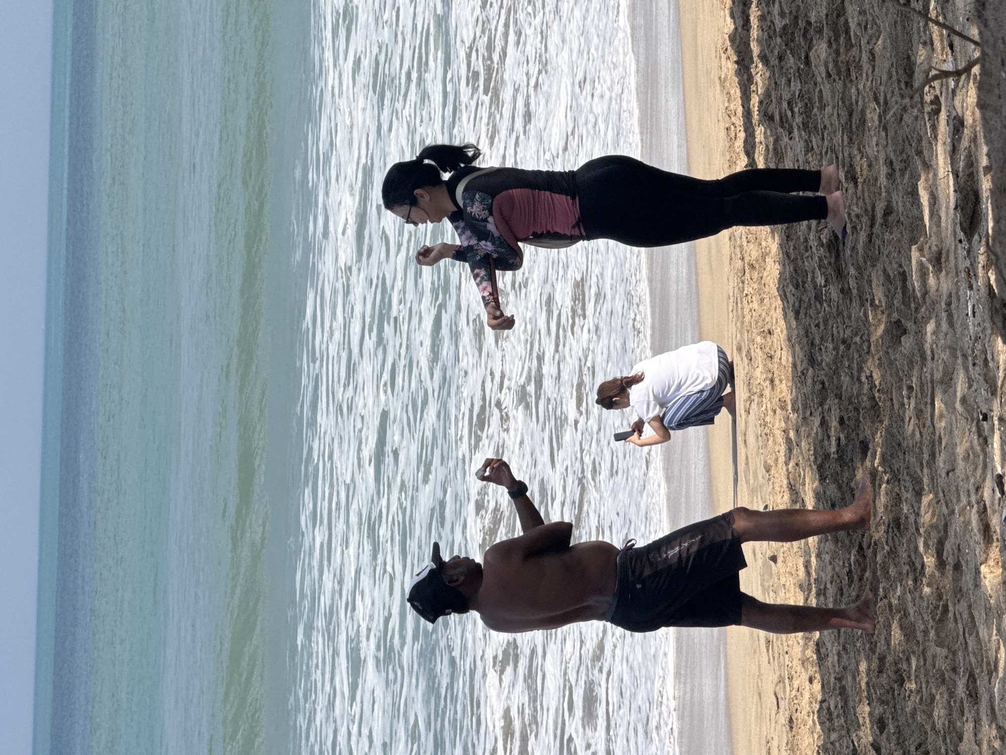 Two adults and one child are on a sandy beach in Kuta, Indonesia, with the ocean in the background. In the foreground, a shirtless man wearing a black baseball cap and dark shorts stands with his arms outstretched, holding what appears to be a small white object in his right hand. To his right, a woman in a long-sleeved rash guard with a floral pattern and black leggings stands with her arms raised, as if stretching. Further back and between the two adults, a child in a white shirt and striped pants is crouching, looking at a smartphone. The water is light blue and waves are breaking on the shore. The sand is light brown and appears to be damp in some areas.