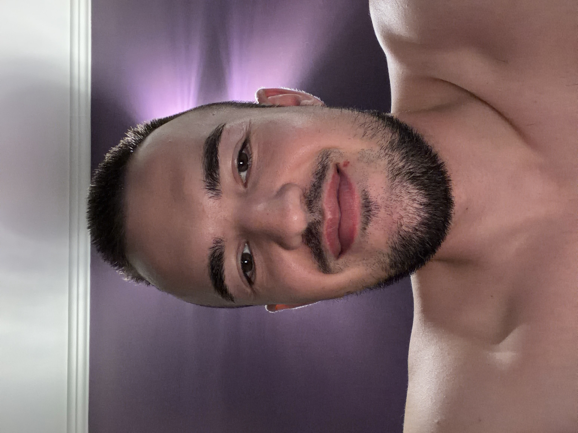 This is a selfie of a fair-skinned man with short, dark hair and a beard. He is wearing a red lipstick. The background is a purple wall with a white molding at the top. The lighting suggests artificial light is being used, possibly from a lamp or overhead fixture, creating a halo effect around his head. The photograph appears to have been taken indoors. There is no identifiable location in the city of Sofia, Bulgaria.