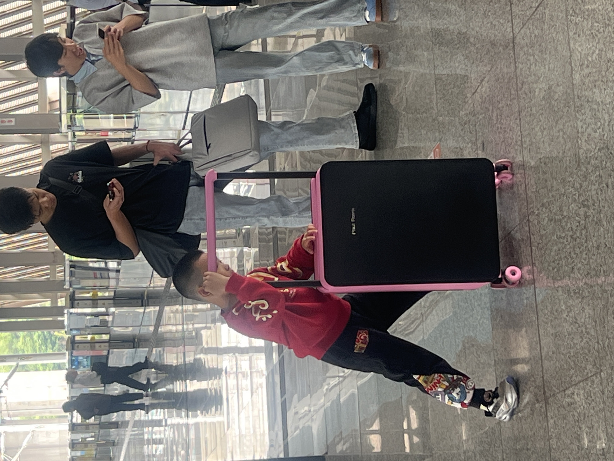 A medium shot shows a boy in a red jacket and black pants holding the handle of a suitcase with a pink frame and black body. A man in a black shirt pulls the suitcase. Another man in a gray blazer and light blue shirt is in the background. The location is Jiangjin, China, in an airport or train station. The floor is gray.