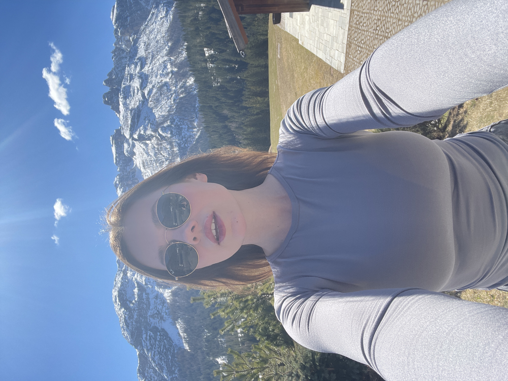 A selfie of a woman wearing sunglasses in Sappada, Italy. The woman is wearing a gray long-sleeved shirt. Snow-covered mountains are visible in the background, along with trees and a blue sky with scattered clouds. Part of a building is visible on the right side of the frame.