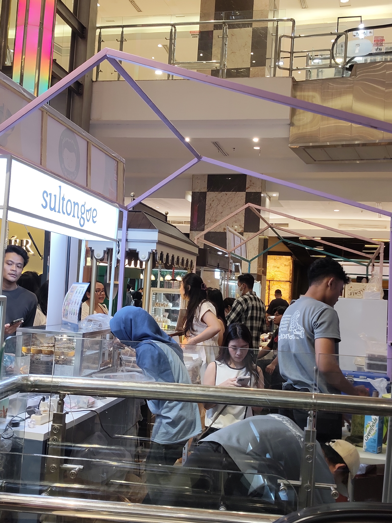The image depicts a bustling scene inside a commercial establishment, possibly a mall or a food court, in the city of Medan, Indonesia. The foreground is dominated by a polished metal railing, suggesting a multi-level indoor space with elevated walkways or viewing areas. Behind the railing, a food stall with a prominent "sultongqe" logo is visible. This stall features a brightly lit sign and a display case containing what appear to be food items, possibly pastries or desserts.
Several individuals are engaged in various activities around the stall. A young man in a grey t-shirt with a graphic print is standing on the right, looking downwards. To his left, a young woman in a white top is holding a smartphone. Further left, a person wearing a blue headscarf and a light blue jacket is attending to items inside the display case. Another woman with glasses, wearing a light-colored top, is visible behind the counter of the stall. Other individuals are seen in the background, including a man in a checkered shirt.
The environment suggests a commercial setting with ambient lighting from overhead fixtures. Decorative structural elements, such as purple and pink geometric frames, are visible above the stall, adding to the visual appeal of the space. The overall impression is one of a lively commercial area where people are browsing and purchasing items.