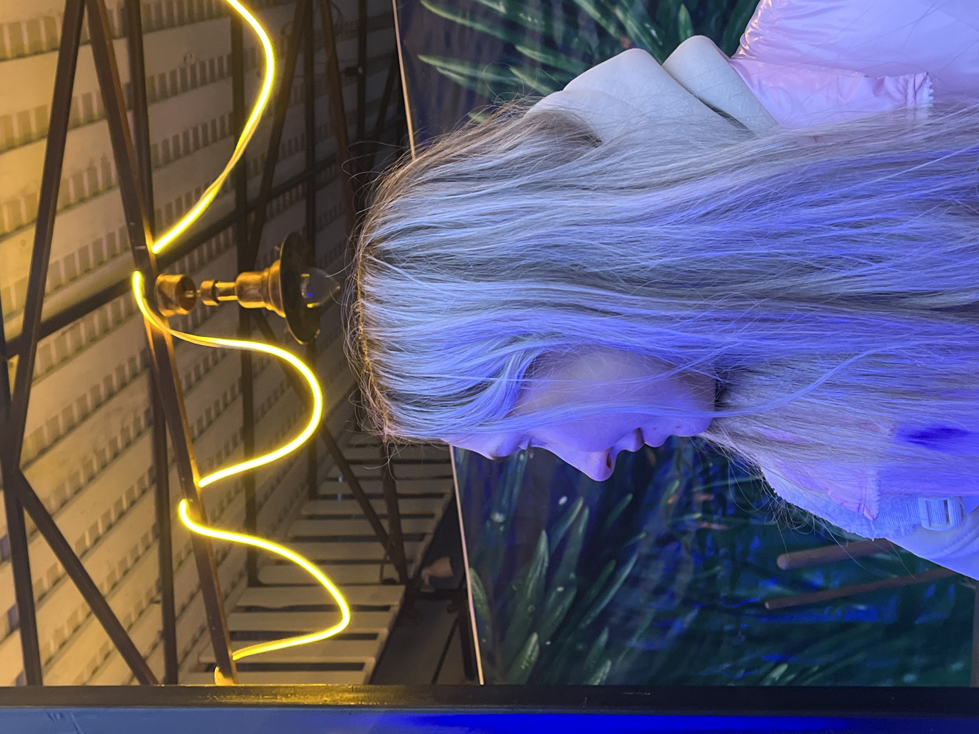 Here is a forensic description of the image:
The image presents a scene featuring a person with long, light blonde hair. The hair appears to be dyed with a blue hue in the lower sections. The person's face is partially visible in profile, with focus on the nose and mouth area. They seem to be wearing light-colored clothing.
In the background, there's a combination of architectural elements and a naturalistic backdrop. On the left side, a structural framework with geometric patterns is visible, illuminated by a winding yellow light fixture. Behind the person, a blurry backdrop suggestive of a lush, green environment is noticeable. The composition suggests an indoor setting with deliberate placement of light, human subject and environmental background. The location is unknown.