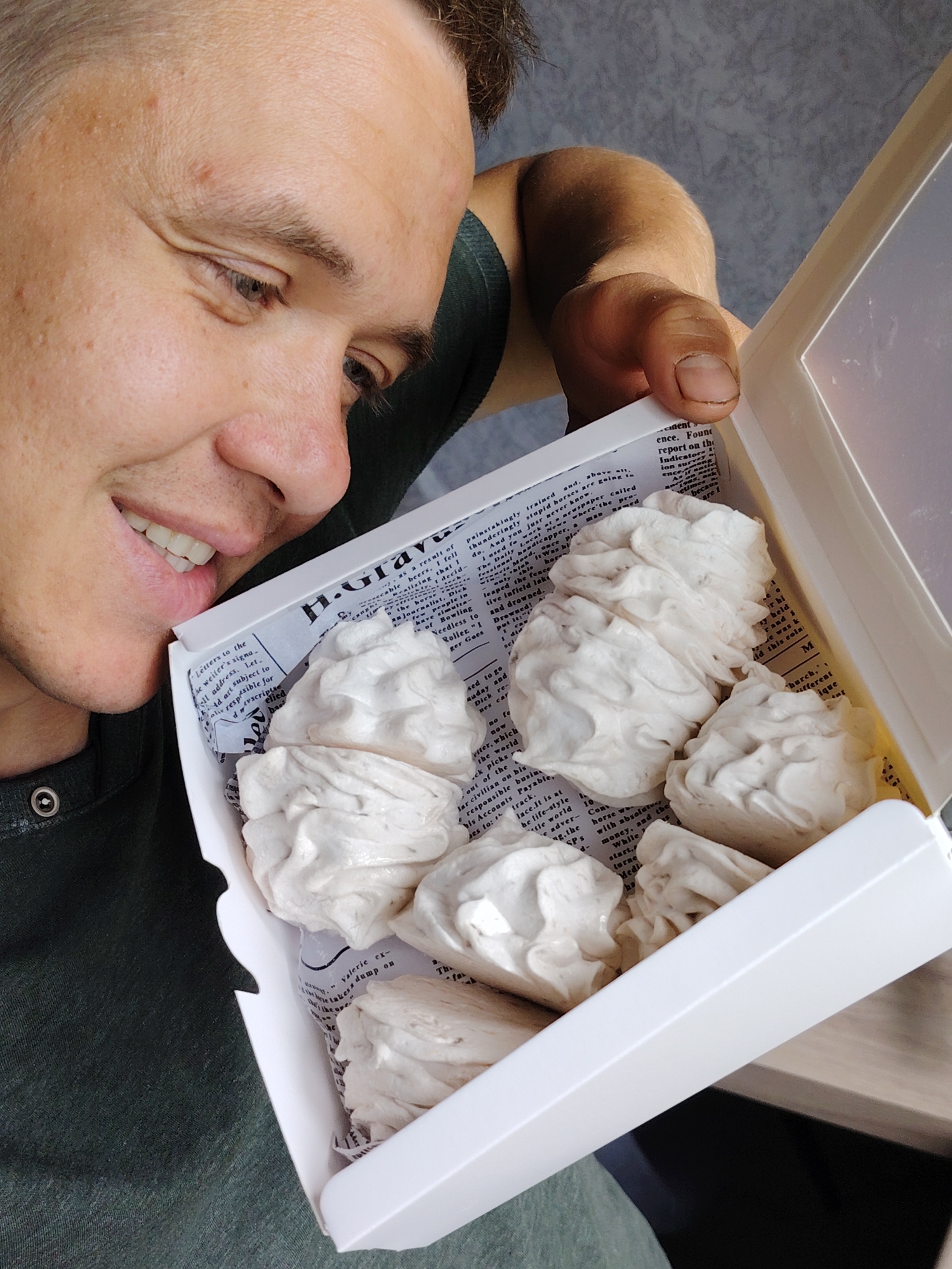 A man, seen from the chest up, smiles as he looks down into a white rectangular box filled with confectionery. He has short brown hair and fair skin, and is wearing a dark green shirt. His right hand, with a short fingernail, is visible holding open the clear plastic lid of the box.
Inside the box are several pieces of white, irregularly shaped, soft-looking confectionery, resembling marshmallows or meringues (zefir). They are arranged on a bed of paper that lines the bottom of the box. This paper is printed with text in a style resembling old newspaper or book pages. Visible text fragments include phrases such as "Letters to the," "Daily Local News," "journalist, Dick," "report on the Indicators," "money," "horse," and "stupid horses."
The man's expression conveys happiness and contentment as he views the treats. The background is a plain light grey wall, with a wooden surface partially visible on the right. The scene appears to be an indoor, daytime setting. This moment takes place in Chaikovsky, Russia.