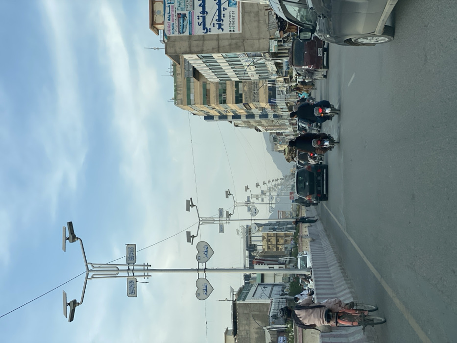 The image depicts a bustling urban street scene in Kandahar, Afghanistan, observed from a low angle. The sky is partly cloudy with light blue patches. The foreground features an asphalt road with visible lane markings.
On the left, a person dressed in light-colored traditional Afghan attire and a dark turban rides a red bicycle, traveling away from the viewer. They are positioned alongside a concrete median barrier that separates the road. In the central lanes, two men on motorcycles, also traveling away from the camera, are prominent. Further down the road, a black Toyota Yaris hatchback is visible, followed by numerous other vehicles and pedestrians.
The street is flanked by multi-story buildings of varying heights and designs, suggesting a commercial and residential district. On the right side, a building displays a large banner with Arabic script and English text reading "HUZAIFA ADVERTISING," alongside contact numbers.
A series of identical streetlights extends down the center of the road, featuring solar panels mounted on their upper arms. Each streetlight includes decorative elements, such as heart-shaped signs bearing Arabic script, and additional rectangular signs with more Arabic text. Overhead power lines are visible crisscrossing the sky. In the far background, distant mountains are discernible above the urban sprawl.