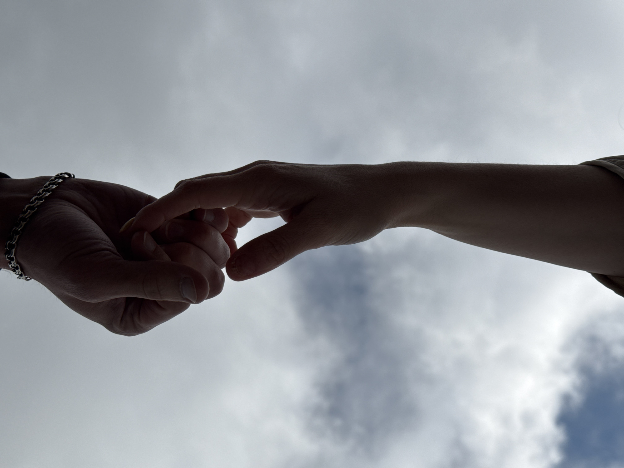 Two hands, one wearing a bracelet, are reaching towards each other in front of a cloudy sky. The location is Chaikovsky, Russia. No specific objects or interactions can be identified.