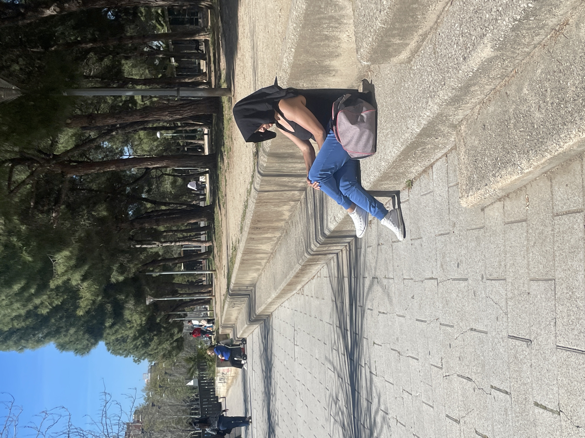 The image shows a person dressed in blue jeans, white shoes, and a dark hooded top sitting on a low concrete wall. A grey backpack rests on the wall next to them. The setting appears to be a park or a pedestrian walkway with a paved surface. To the left are trees and other individuals can be seen in the distance. The location is Sant Adrià de Besòs, Spain, based on visual features consistent with parks found in that area.
