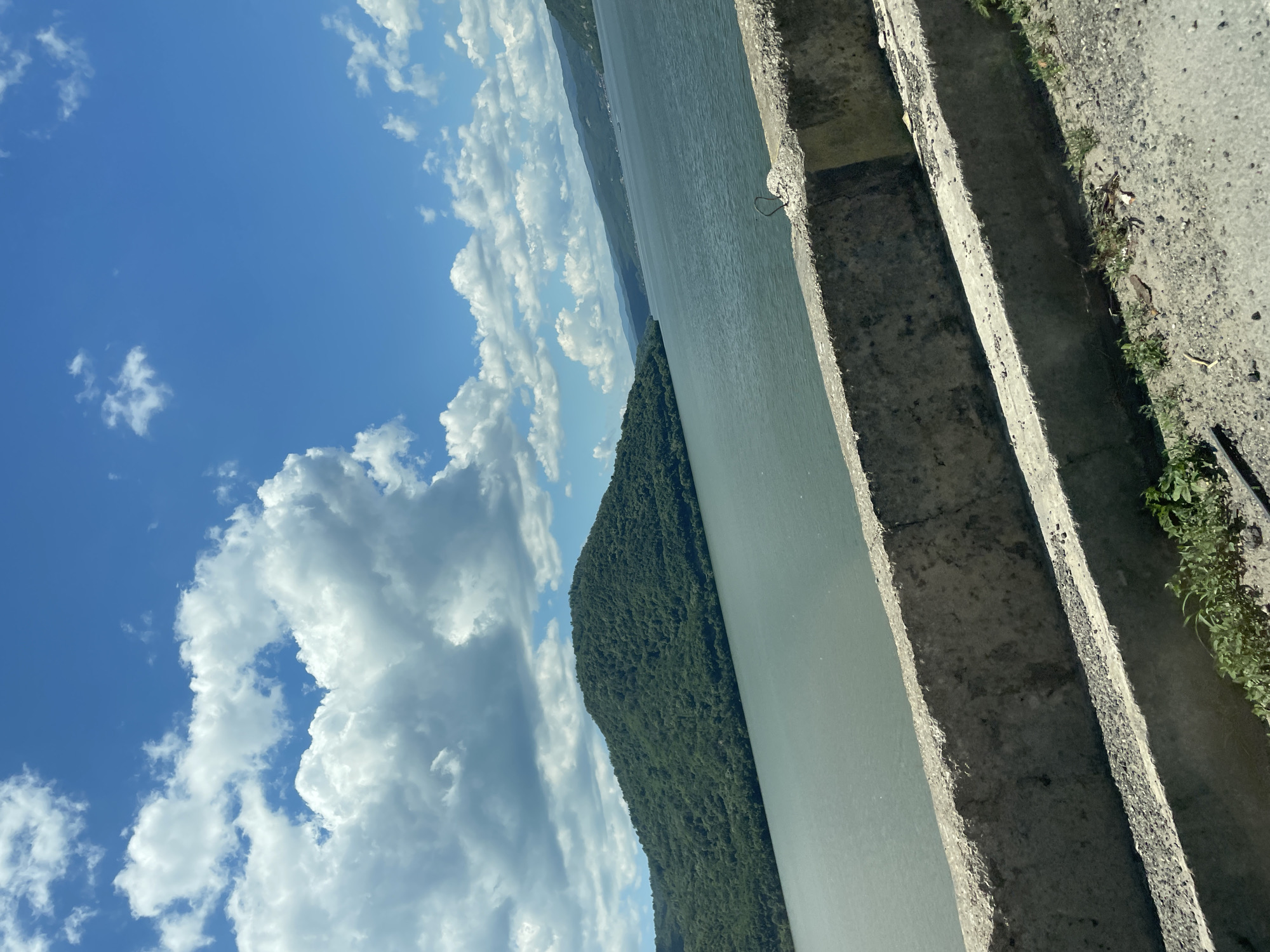 The image presents a daytime landscape capturing a wide body of water, identified as the Danube River, within the geographical vicinity of Drobeta-Turnu Severin, Romania. The upper portion of the frame is dominated by a bright blue sky partially filled with numerous white cumulus clouds of various sizes and formations.
In the immediate foreground, a weathered concrete barrier, appearing as a multi-tiered guardrail or embankment, extends horizontally across the lower part of the image. This structure exhibits a coarse, granular texture and is grey in coloration. Below the concrete barrier, a narrow strip of ground composed of dirt and gravel is visible, supporting sparse patches of green vegetation and some minor debris.
Beyond the concrete barrier, a substantial expanse of water occupies the middle ground. The water surface displays a muted greenish-grey tone and appears relatively calm. To the left, a large, steep landmass, completely covered in dense, dark green deciduous forest, rises directly from the water's edge. On the right side of the water body, several receding, forested hills or mountains are visible in the distance, extending towards the horizon under the cloud-strewn sky.
No human subjects, animals, or man-made structures other than the concrete barrier and distant hints of settlement on the far shore are discernible within the frame. The lighting suggests direct sunlight from a clear sky.