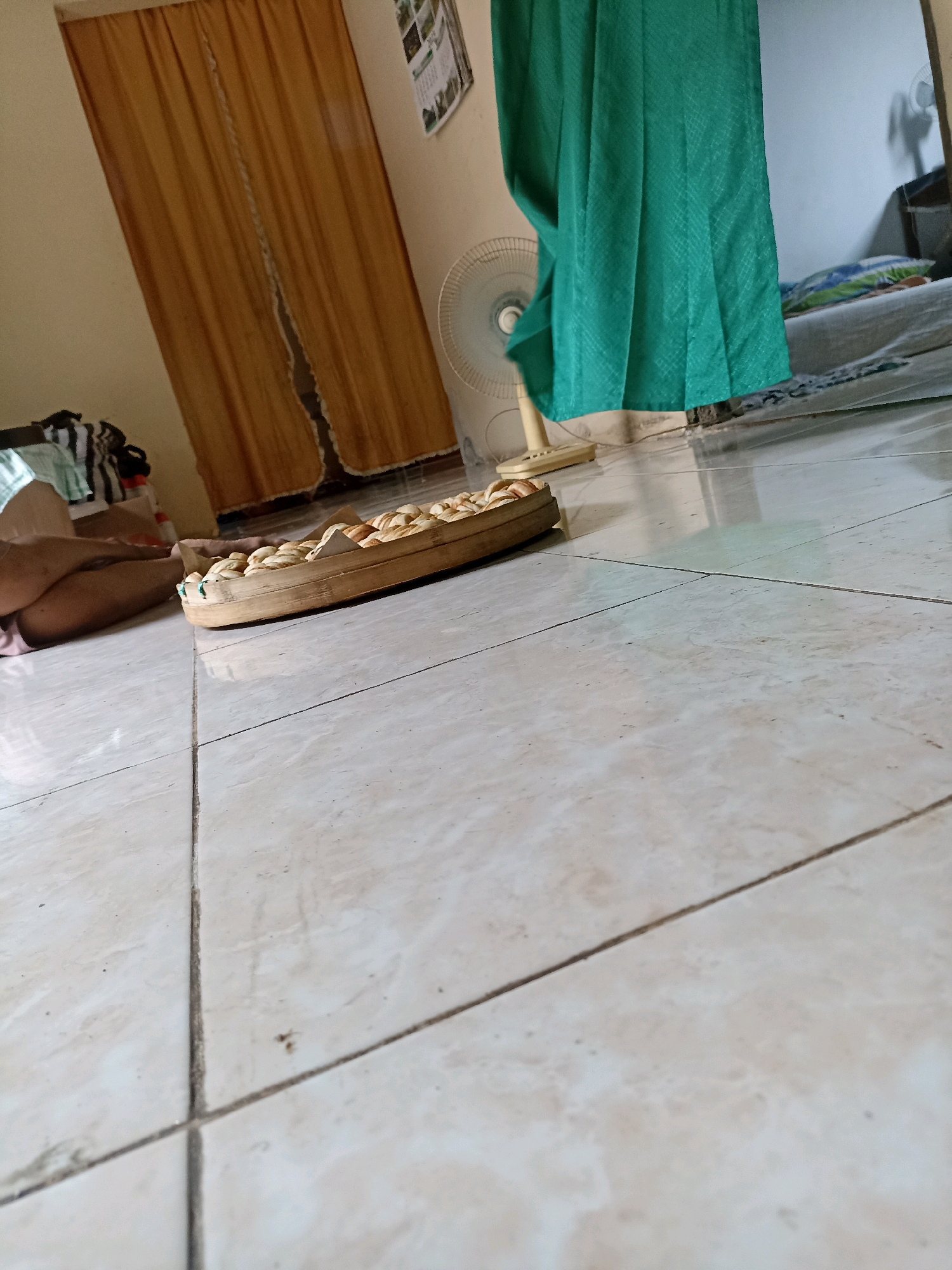 The image depicts an indoor scene in a tiled room, likely a residential dwelling in Sonobekel, Indonesia. In the foreground, a person's legs are visible, resting on the floor next to a circular woven tray filled with what appear to be dried snacks or food items. The tray is placed on the tiled floor. In the background, a standing fan is visible, partially obscured by a green curtain. To the left, a golden-brown curtain is drawn, revealing some stacked clothing or fabric behind it. Further in the background, a bed with pillows is visible, suggesting a bedroom or living area. The overall environment appears to be domestic and casually arranged.