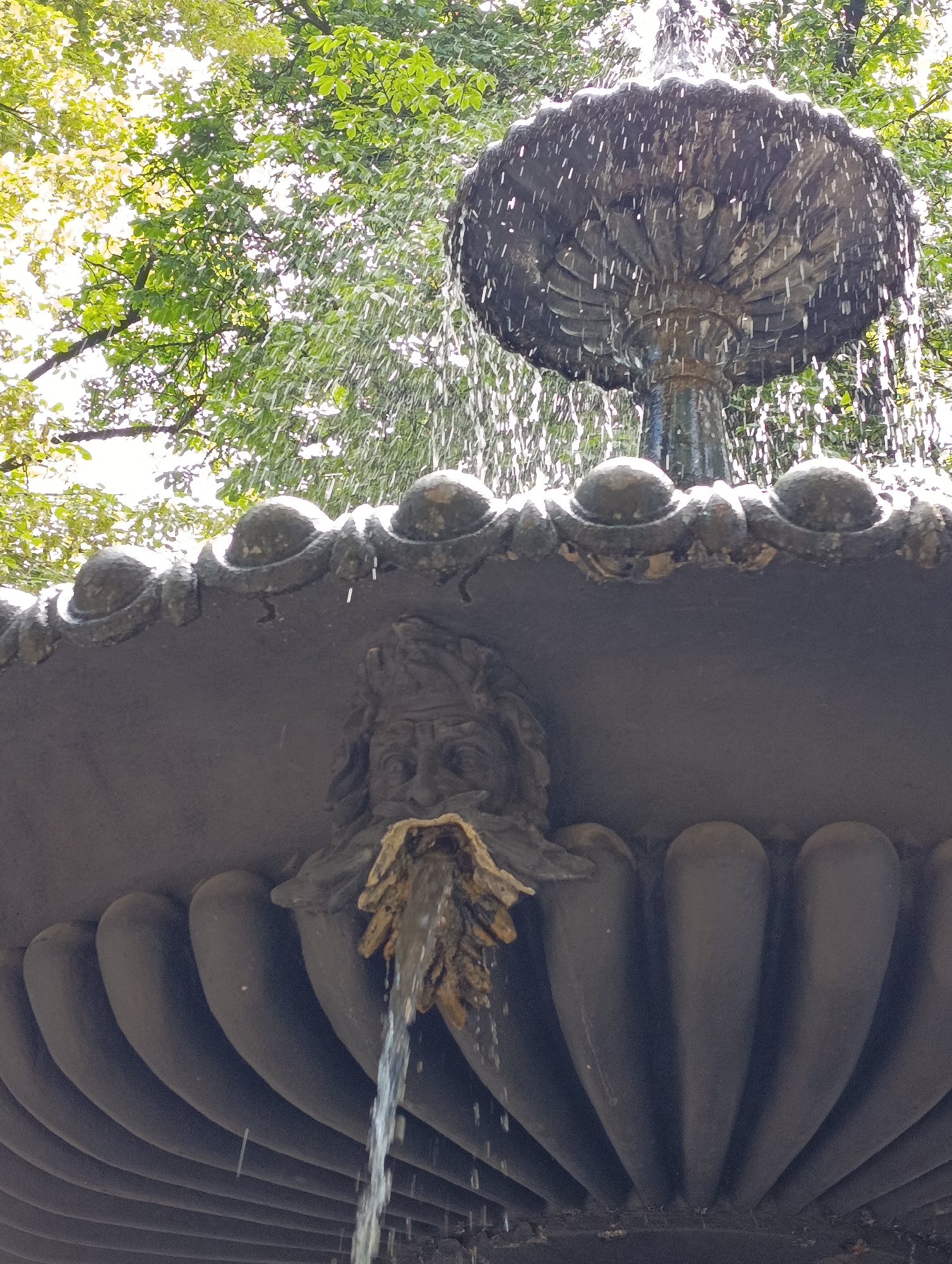 The image shows a close-up of a fountain with water cascading from its top bowl, which has a decorative, petal-like structure. Below the top bowl, a sculpted face is integrated into the fountain's basin, with water flowing from its mouth area, which appears to be a leaf-like ornament. The fountain is set against a backdrop of lush green trees and bright sky, suggesting an outdoor location, likely in a park or garden. The fountain itself appears to be made of stone or a similar material and has an ornate, classical design. The image was taken in Kyiv, Ukraine.