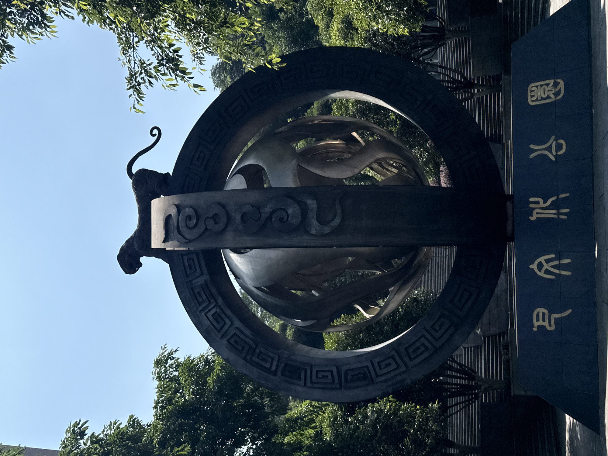 The image captures a grand outdoor monument or sculpture under a clear blue sky, framed by lush green trees and foliage. The central feature is a dark-colored, multi-layered structure dominated by large circular elements. The outermost ring and an inner one are adorned with intricate, repeating geometric patterns, resembling meanders or key patterns. Within these circular frames, abstract, flowing sculptural elements, possibly in a lighter metallic tone, are visible, suggesting organic forms like waves or clouds.
Perched on the top-left of the main structure is a stylized figure of what appears to be a mythological creature or animal, featuring a distinct head and a long, elegantly curled tail.
To the right of the monument, a dark blue vertical slab or wall is prominent. This slab bears three gold-colored Chinese characters arranged vertically. Reading from top to bottom, the characters are "巴南园" (Bā Nán Yuán), which translates to "Banan Park" or "Banan Garden," indicating the location.
The scene is illuminated by bright daylight, suggested by the clear blue sky and the lighting on the trees and monument. No people are visible, maintaining a focus on the monument's artistic and cultural details within its natural setting. The overall impression is one of a significant public artwork or landmark.