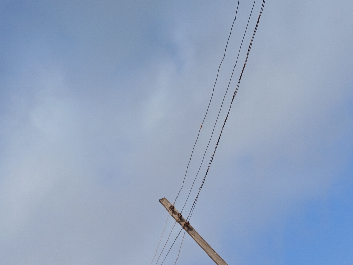The scene, located in Adikpo, Nigeria, captures an upward view of the sky. The majority of the frame is occupied by a light blue sky with diffuse white clouds scattered throughout. In the lower-right quadrant, a weathered wooden utility pole is visible, extending diagonally upwards and towards the left. Three dark-colored electrical or communication wires are strung parallel to each other, attached to the visible portion of the pole, and traverse the sky from the lower-left to the upper-left section of the image. There are no people, explicit actions, or additional significant objects visible within the frame.