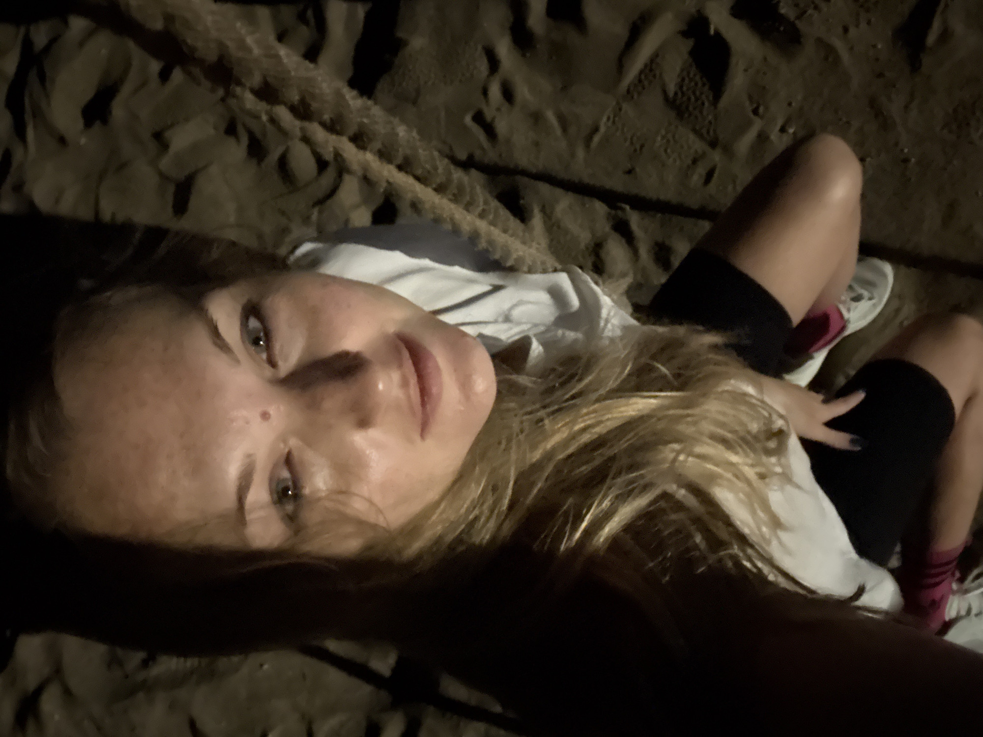 A high-angle, close-up selfie taken at night shows a woman with blonde hair and fair skin, wearing a white top and black shorts. She is kneeling on sandy ground, with a thick rope running diagonally across the image to her right. Her face is partially illuminated by a light source from above, casting shadows. The background appears to be a beach with footprints in the sand. The scene is dark, suggesting an outdoor setting at night, possibly on a beach in Denpasar, Indonesia.