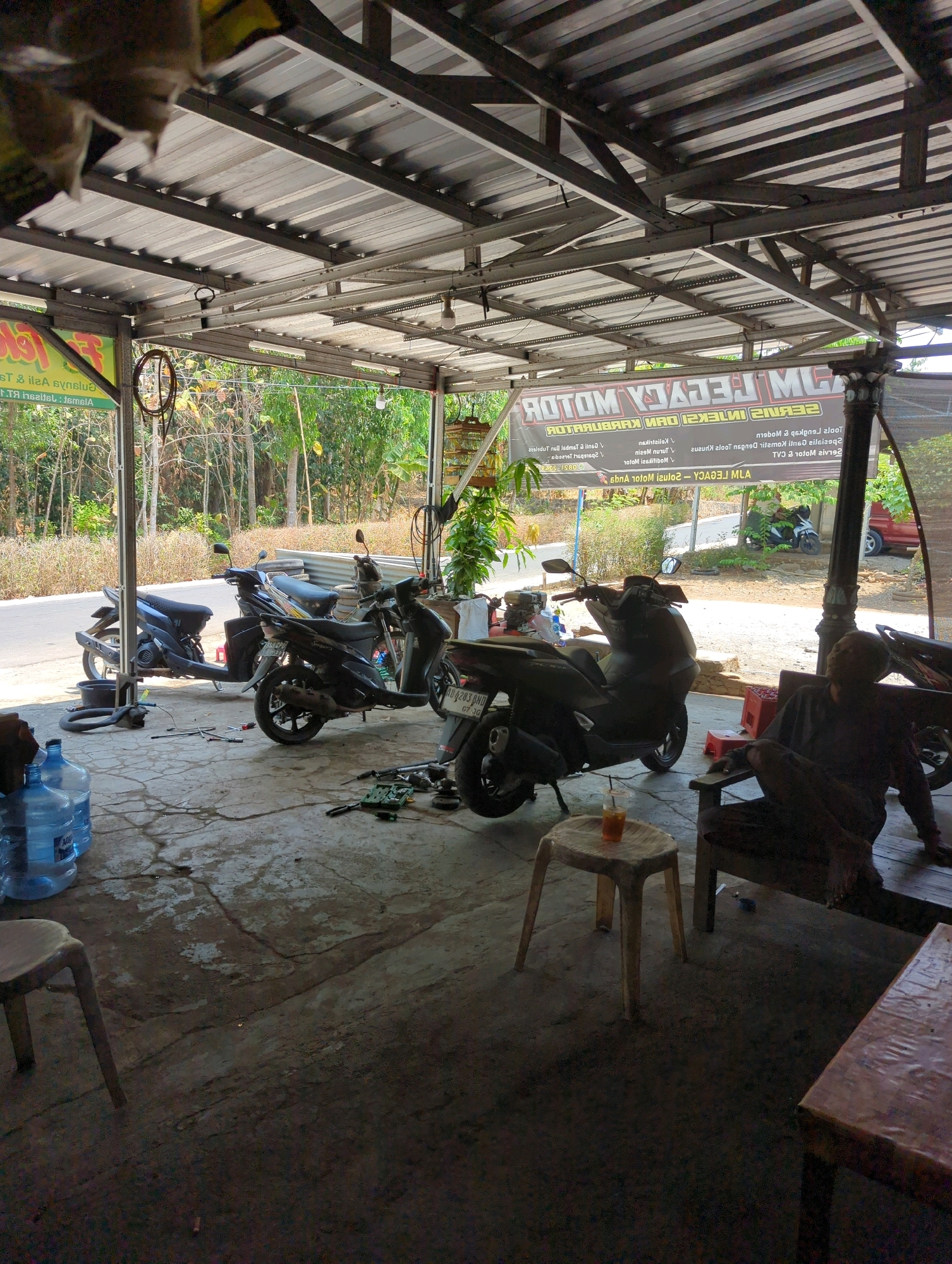 The image depicts an outdoor motorcycle repair shop in Kedungdowo, Indonesia. The scene is covered by a corrugated metal roof supported by metal beams and pillars. In the foreground, on the left, are two large blue water jugs. In the center, several motorcycles are parked, including a black scooter on the left and a darker scooter in the middle, with tools and parts scattered on the concrete floor around them. A small plastic stool with a drink on it is placed in front of the central scooter. To the right, a person is seated on a wooden bench, partially obscured by shadow. A wooden table is visible in the far right foreground. In the background, a banner with text that appears to be in Indonesian is hung, and beyond that, lush green trees and vegetation are visible, along with a road. A red car is also parked in the background. The overall environment suggests a functional, albeit somewhat cluttered, repair facility.