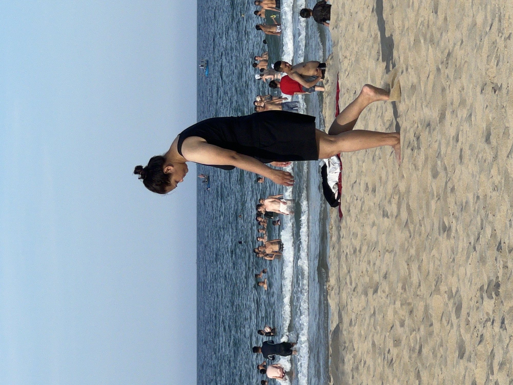 A medium shot captures a woman in a black dress walking on a sandy beach. She is facing towards the right, with her head bowed and her hair pulled up in a bun. Her right foot is mid-stride, kicking up a small amount of sand. In the background, the ocean is visible with numerous people swimming and wading in the water. Waves are breaking on the shore. The sky is a clear blue. The scene appears to be on a beach in Da Nang, Vietnam, given the context of the question.