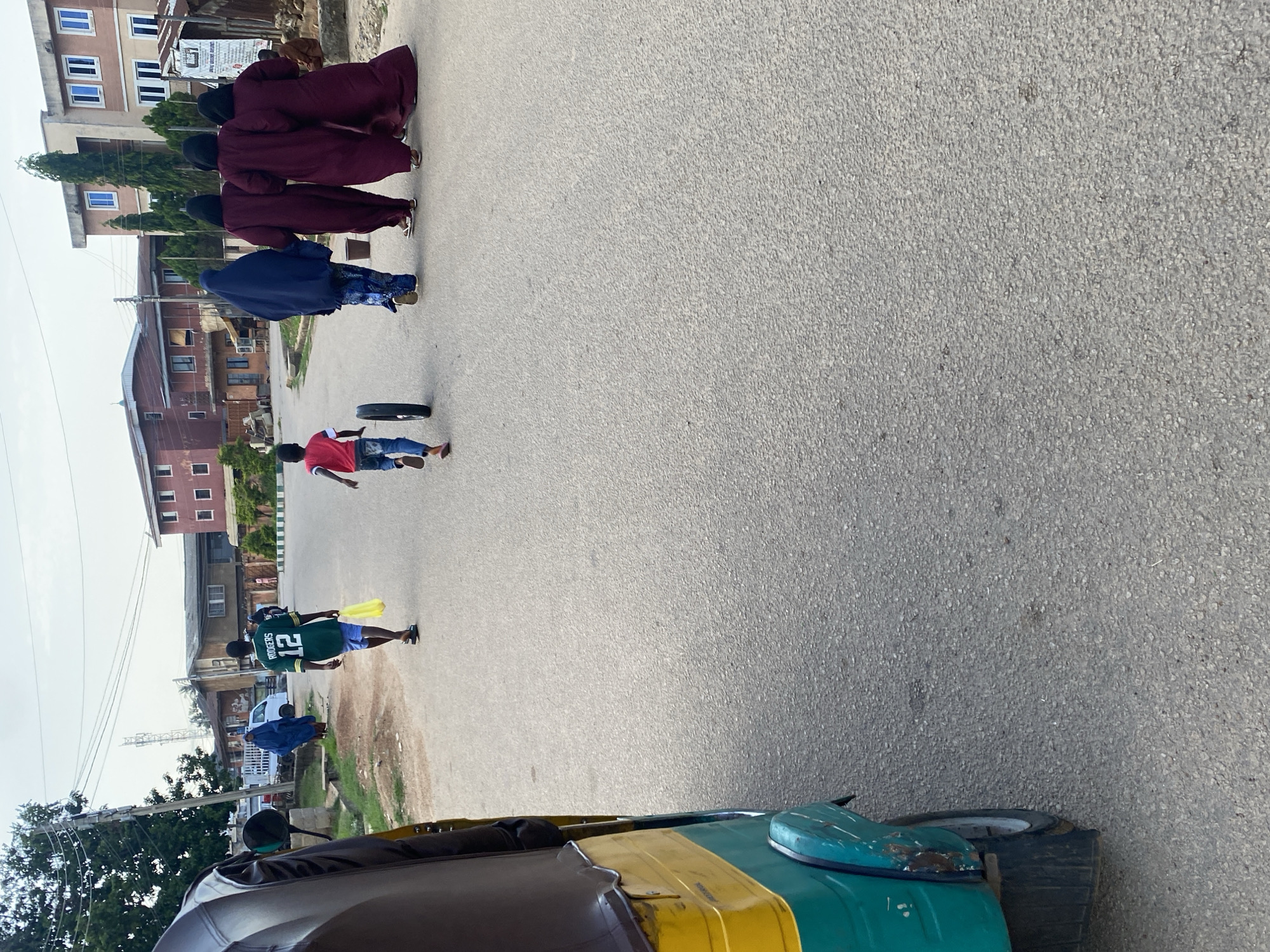 In Jos, Nigeria, an outdoor scene unfolds on an asphalt road. In the foreground, a partial view of a yellow and green autorickshaw is visible on the left. Further down the road, a young boy in a red shirt and blue jeans runs ahead, pushing a spare tire. Behind him, a young man in a green jersey with the number 12 and the name "RODGERS" walks away from the camera, carrying a yellow plastic bag. To the right of the boy, a group of four women dressed in dark red abayas and head coverings walk together. In the background, buildings of various sizes and architectural styles line the street, some with visible windows and balconies. Trees and utility poles add to the urban landscape. The sky above is overcast.