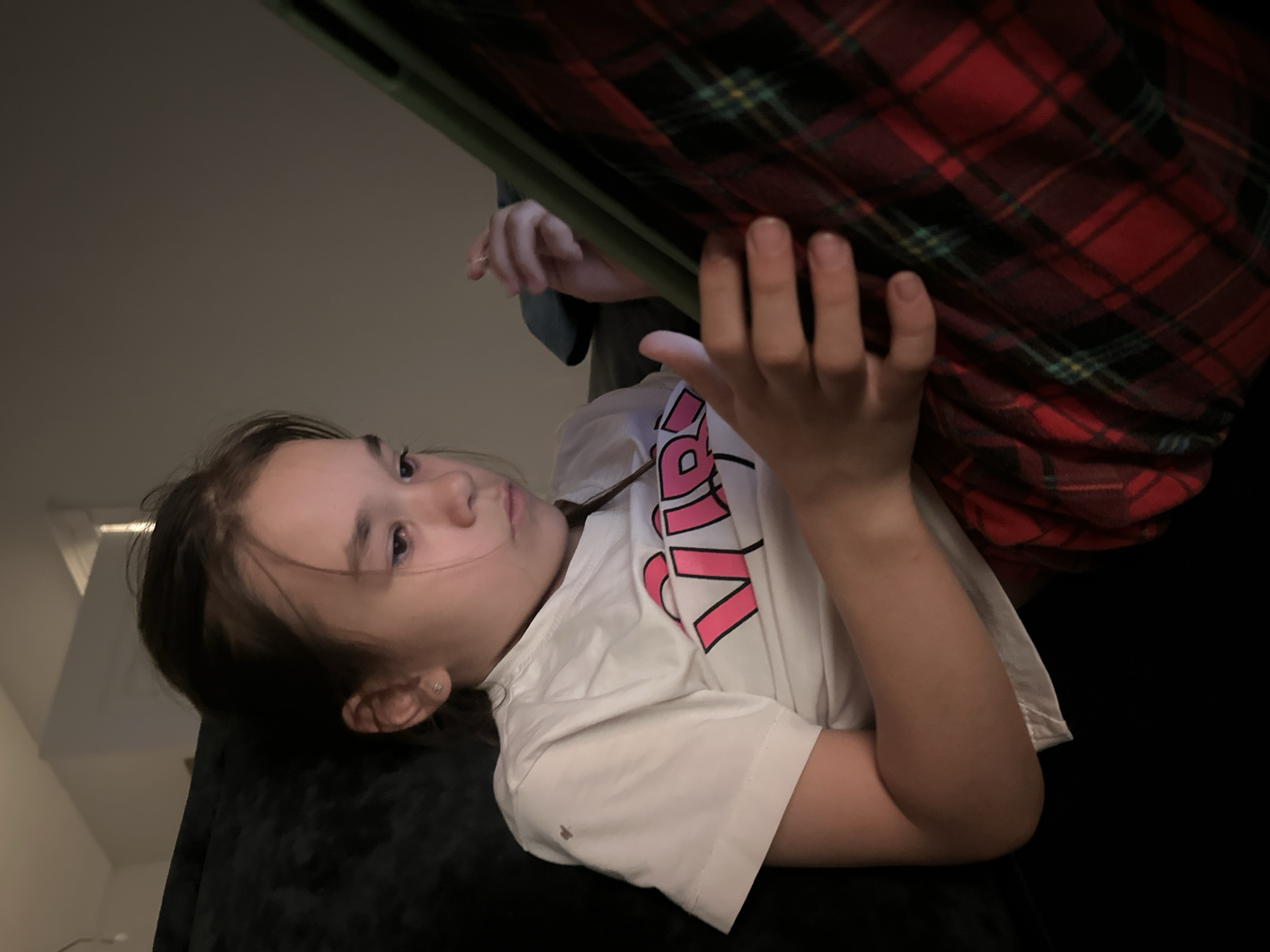 A girl is lying down and holding a tablet. She is wearing a white t-shirt with pink writing. An arm wearing a red and black plaid shirt is above her. The location of the photo is in a dimly lit room. The location in the city of Kharnavy, Russia cannot be determined from this image.