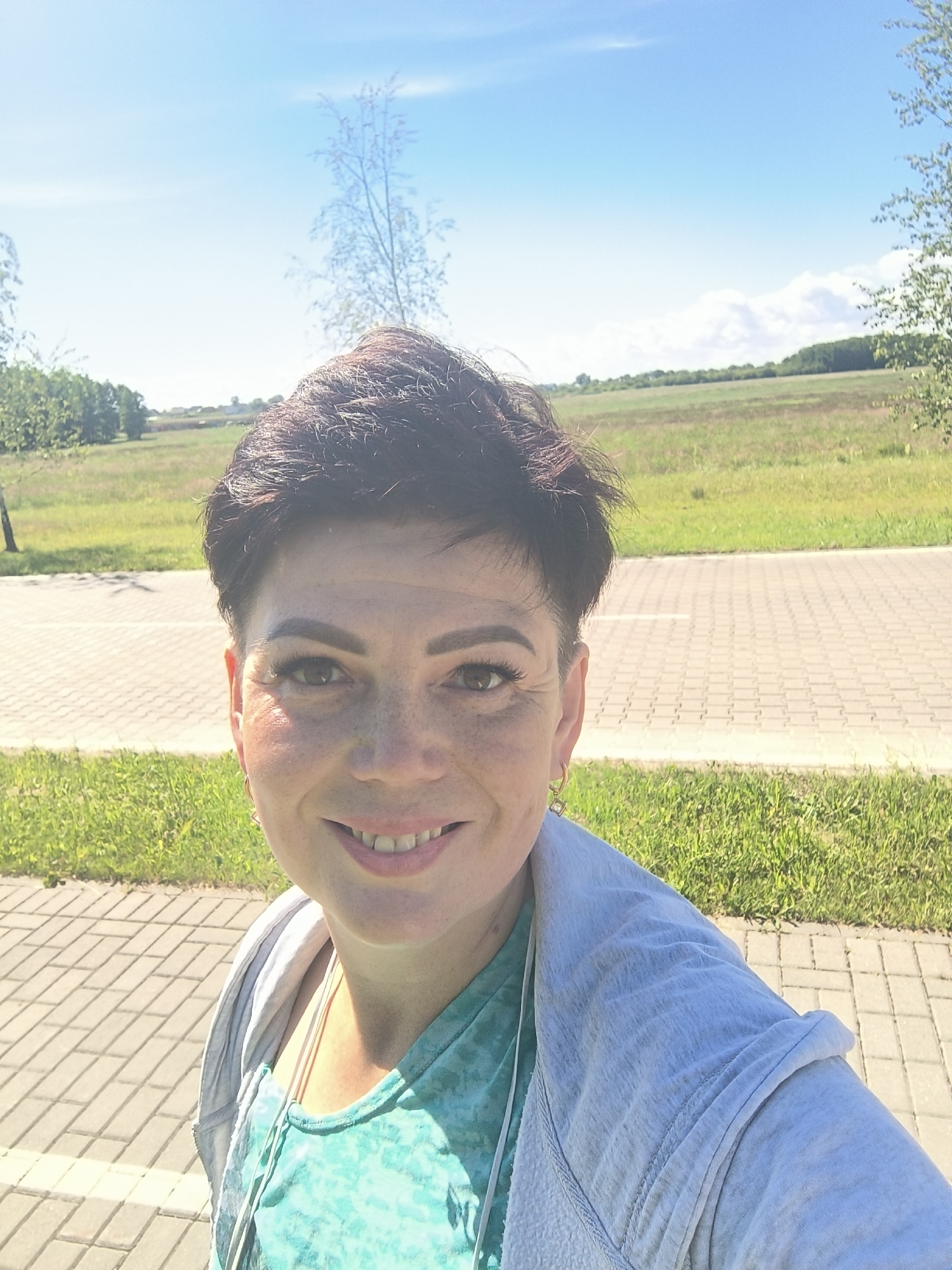 A mid-shot, selfie-style photograph features a woman with short, dark hair, smiling and looking at the camera. She is wearing a turquoise, tie-dye patterned shirt and a light gray hooded jacket. The background consists of a paved walkway, green grass, and a field under a bright blue sky with some scattered clouds. The location is identified as Laskino, Russia. The lighting suggests it is daytime, likely sunny. The woman has freckles and is wearing subtle makeup. A single earring is visible on her right ear. The overall scene depicts a casual outdoor setting.