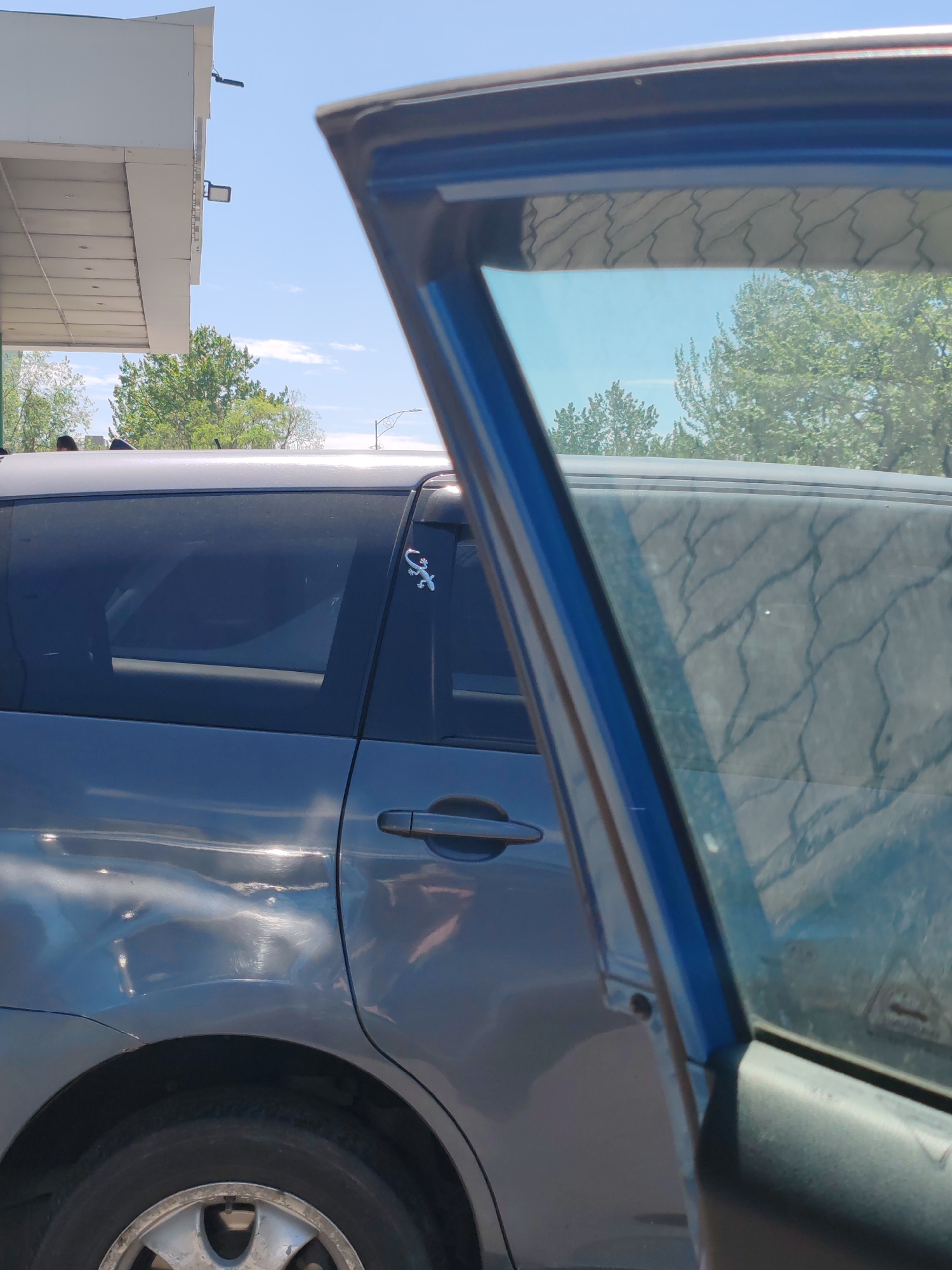 In a sunlit outdoor environment, a grey minivan is parked next to a building with a white awning. The driver's side door is open, revealing the interior. A small, silver gecko sticker is affixed to the window pillar above the rear door. The minivan's body shows noticeable dents and scratches on the rear quarter panel. The sky is blue with scattered white clouds, and green trees are visible in the background. The ground appears to be paved.